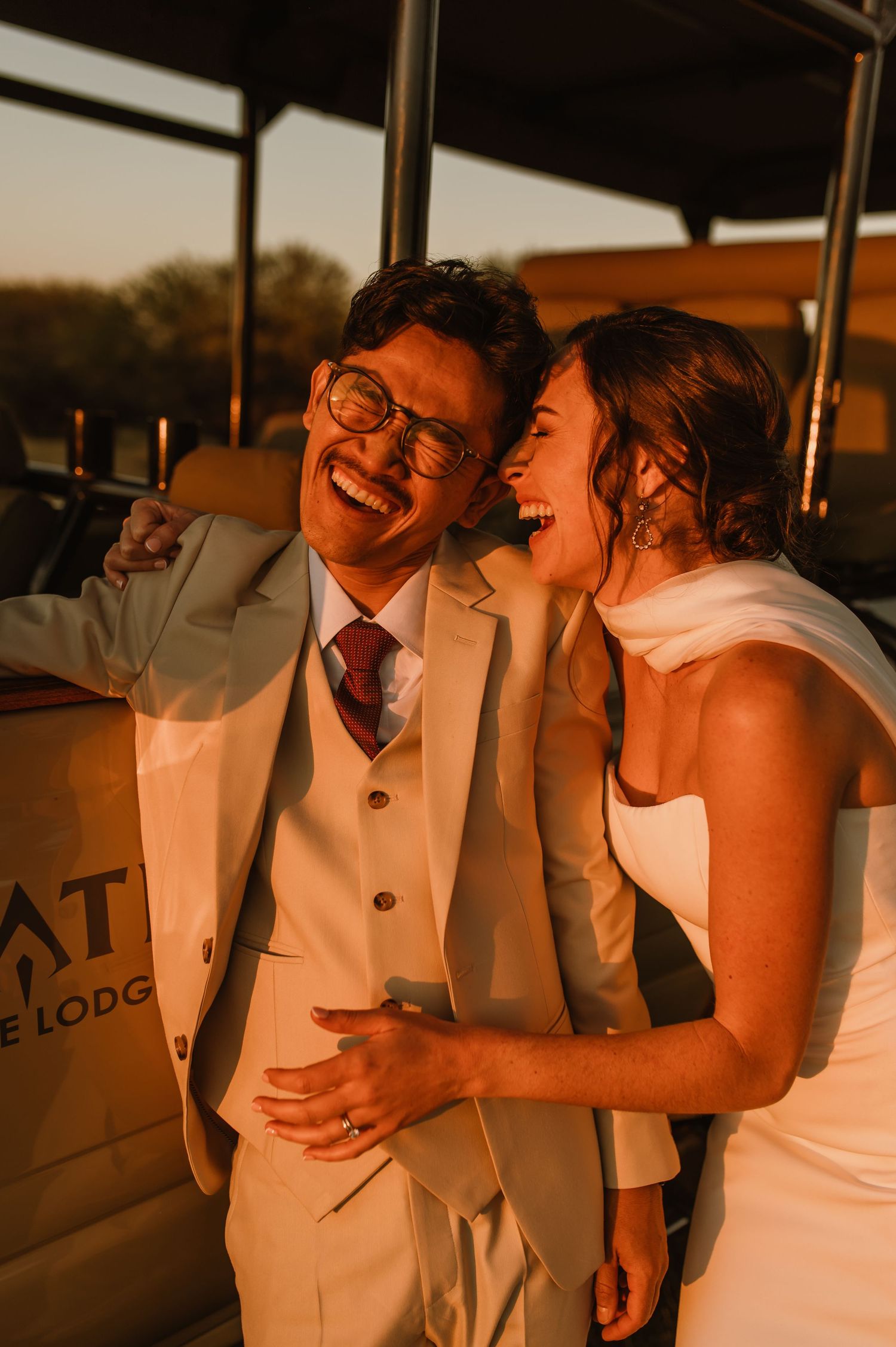 Laughing bride and groom in golden sunset light, wearing wedding attire, sharing a joyful embrace at an outdoor lodge.