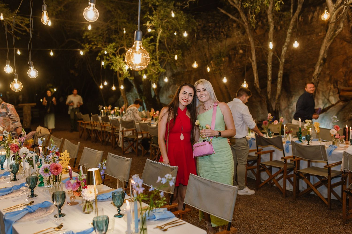 Two smiling women pose at an outdoor wedding reception under warm Edison bulb string lights.
