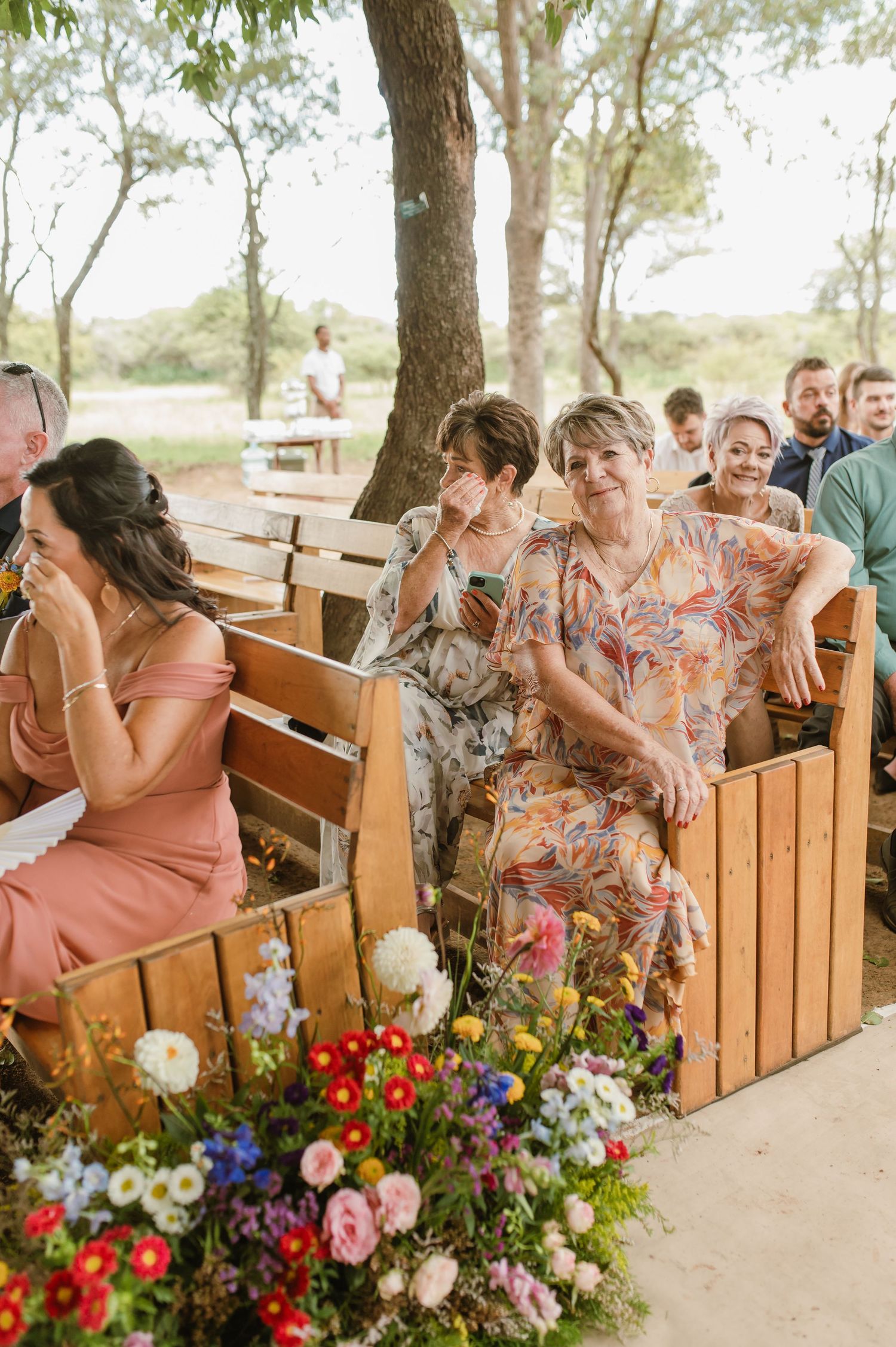 Wedding guests seated on wooden benches at an outdoor ceremony surrounded by trees and colorful floral arrangements.
