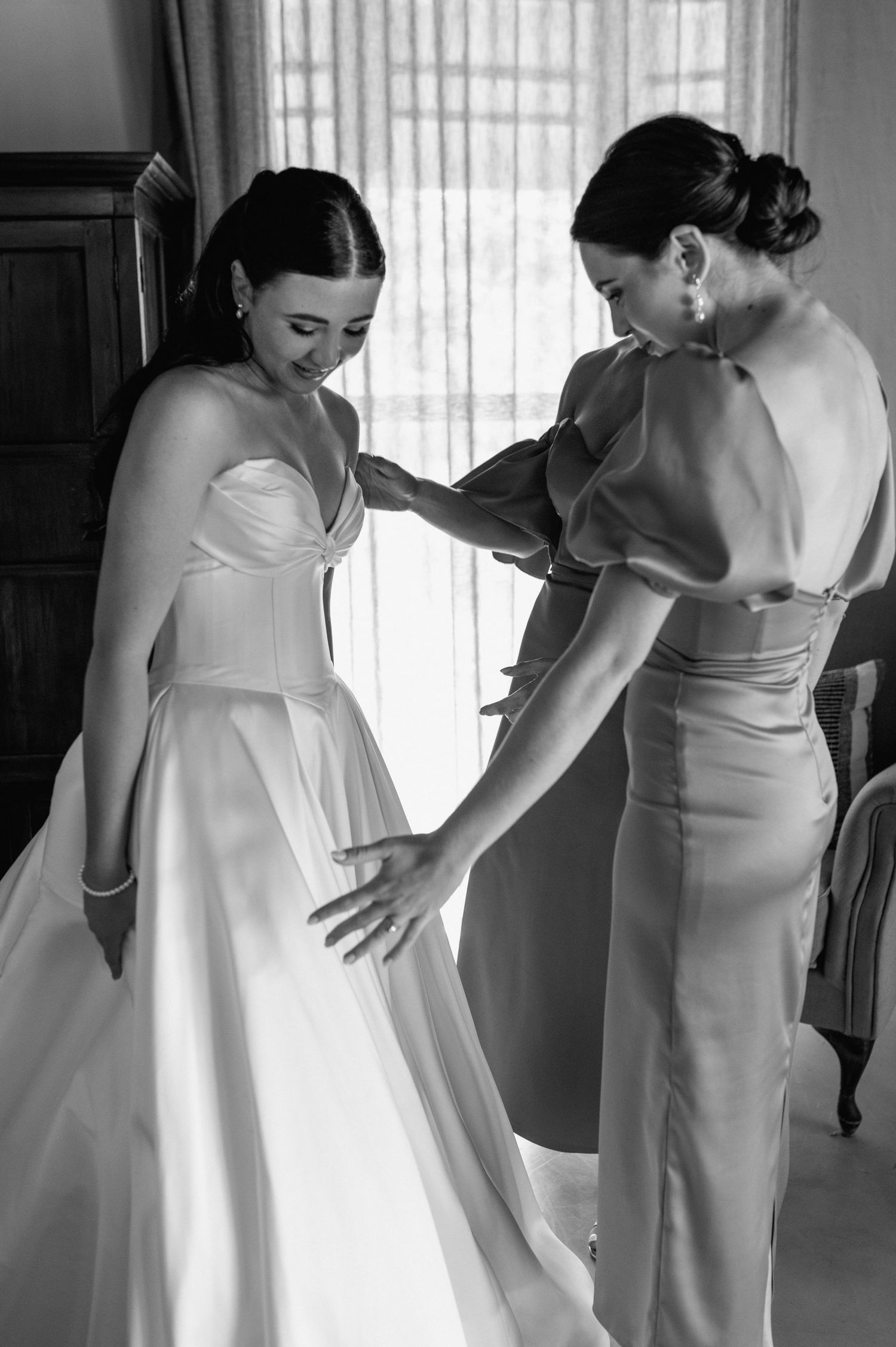 Black and white photo of bride in strapless gown being helped into her wedding dress by bridesmaid.