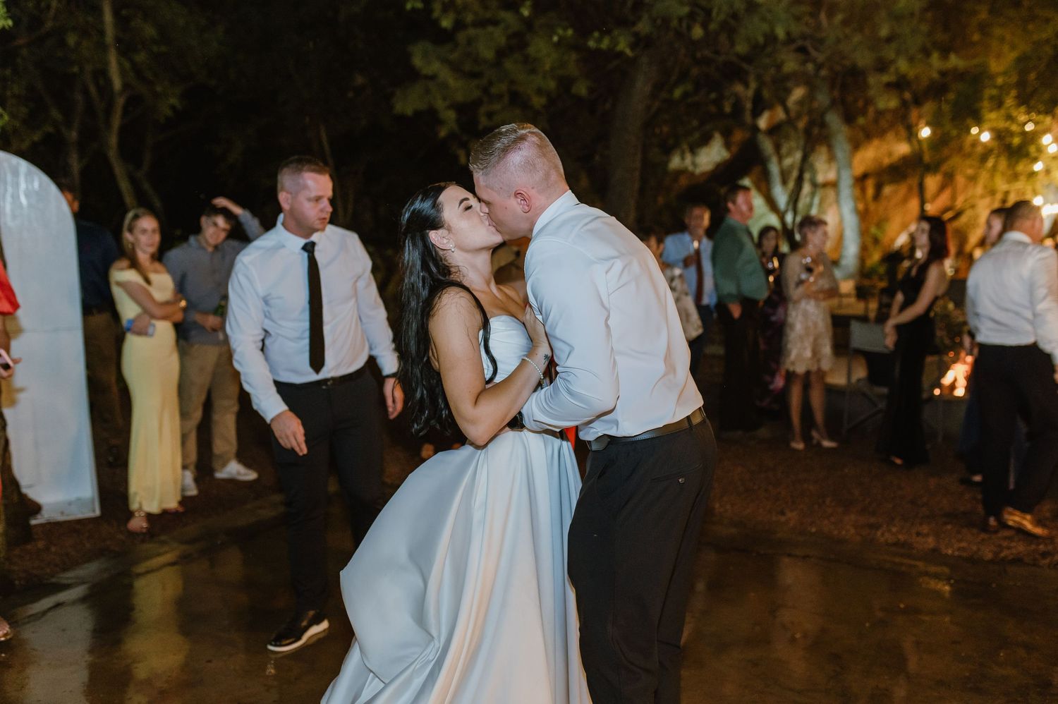 Bride and groom sharing a first dance kiss outdoors at night surrounded by wedding guests under string lights.