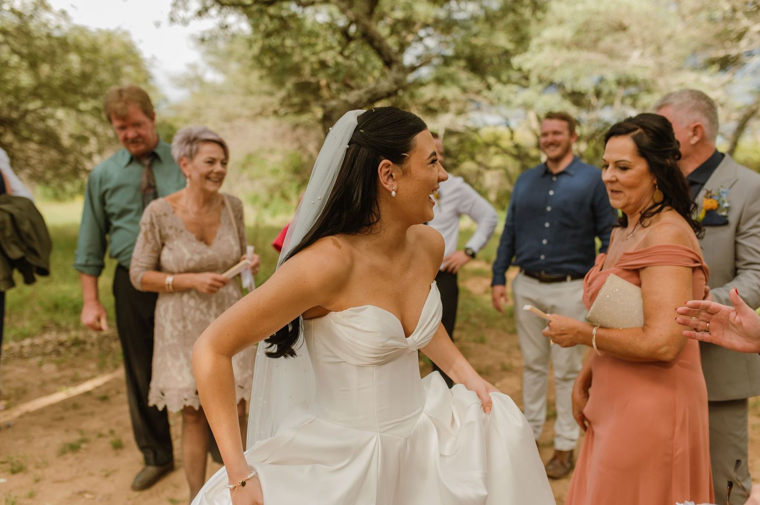 Bride in strapless white gown laughing joyfully with wedding guests after outdoor ceremony.