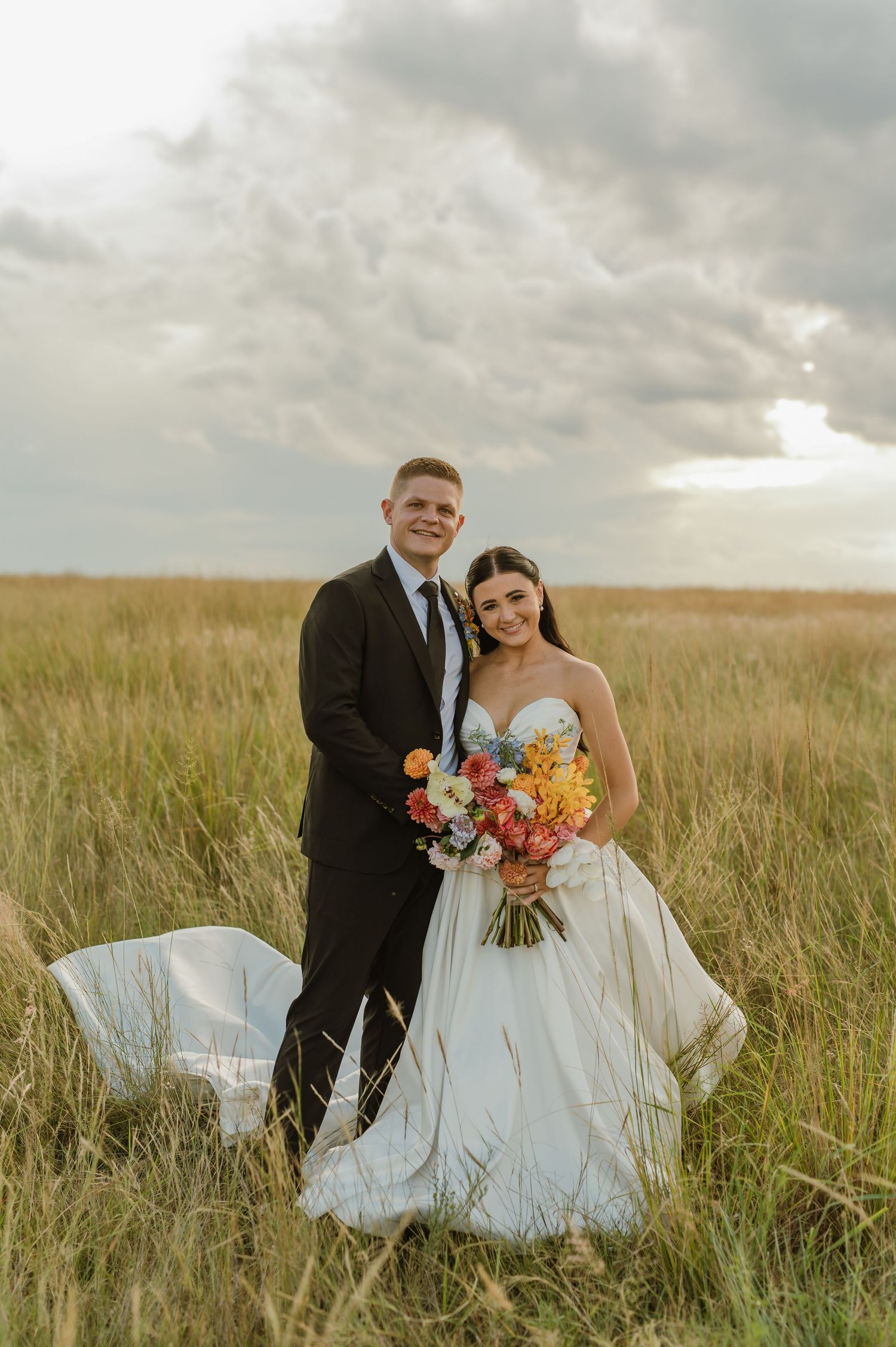 Bride and groom portrait in golden grass field with dramatic cloudy sky and colorful bridal bouquet.