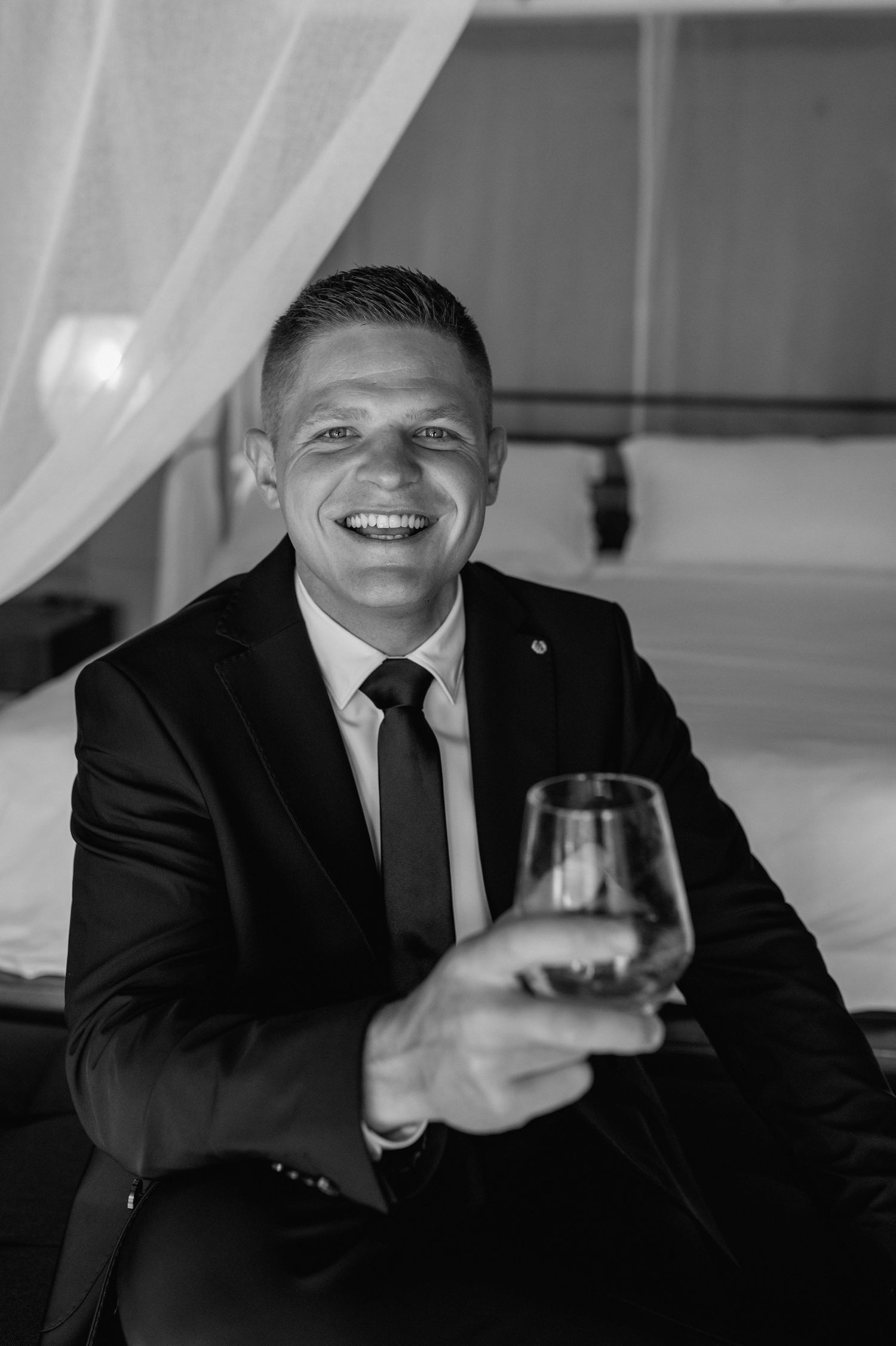 Black and white portrait of a groom in a suit raising a whiskey glass with a confident smile.