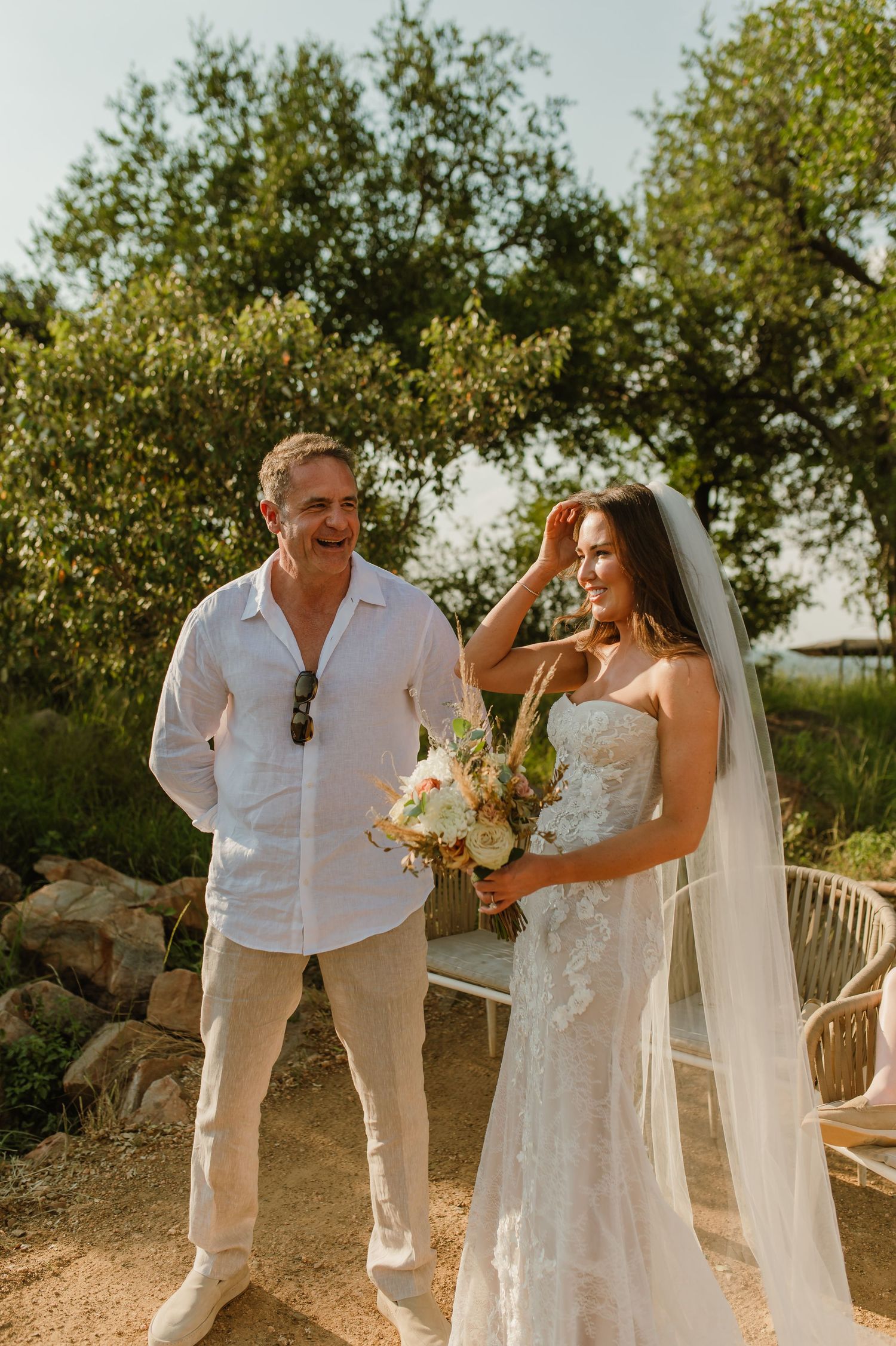 Bride in strapless lace wedding dress and veil laughing with a man in white linen outdoors.