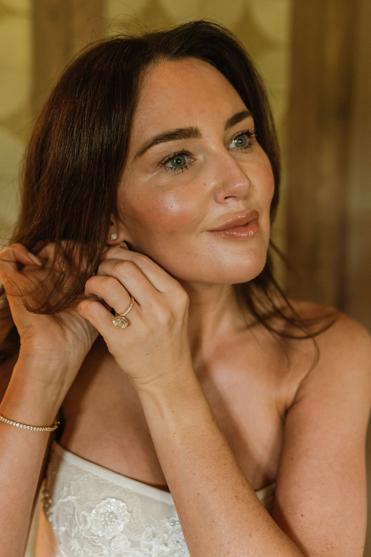 Beautiful bride with green eyes putting on pearl earring in soft golden light before wedding ceremony.