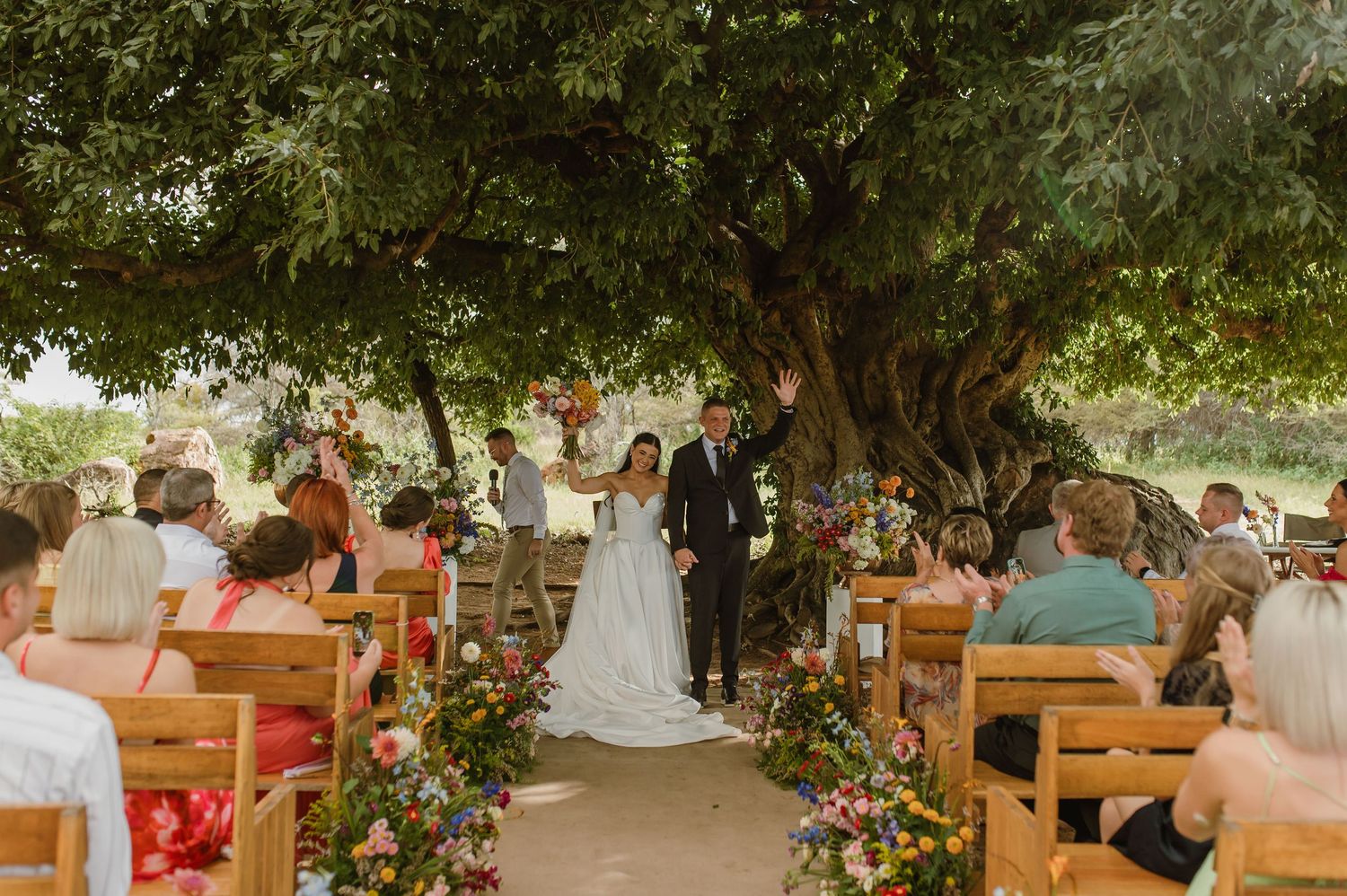 Outdoor wedding ceremony under a large tree with floral aisle and wooden chairs filled with guests.