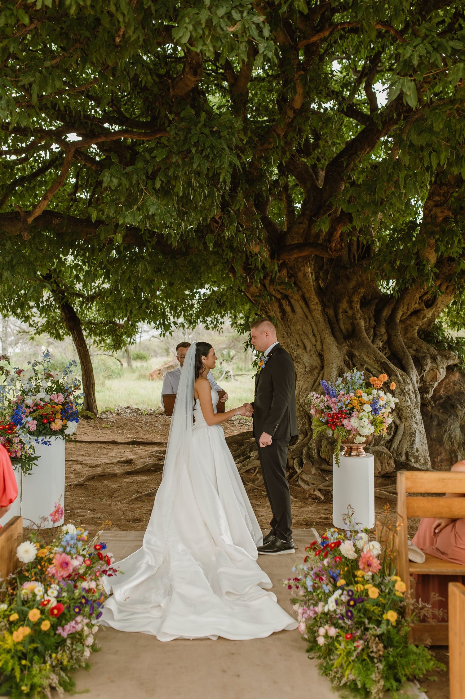 Bride in white gown and veil exchanges vows with groom under large tree adorned with colorful floral arrangements.