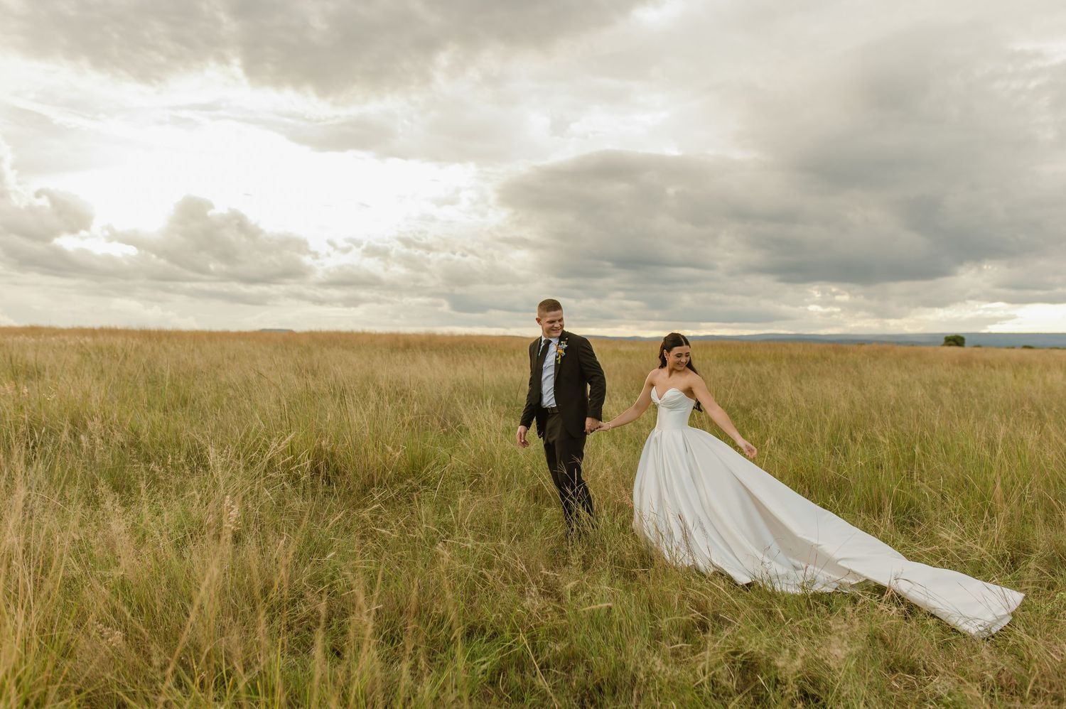 Bride and groom walking hand in hand through a vast open field under dramatic stormy skies.