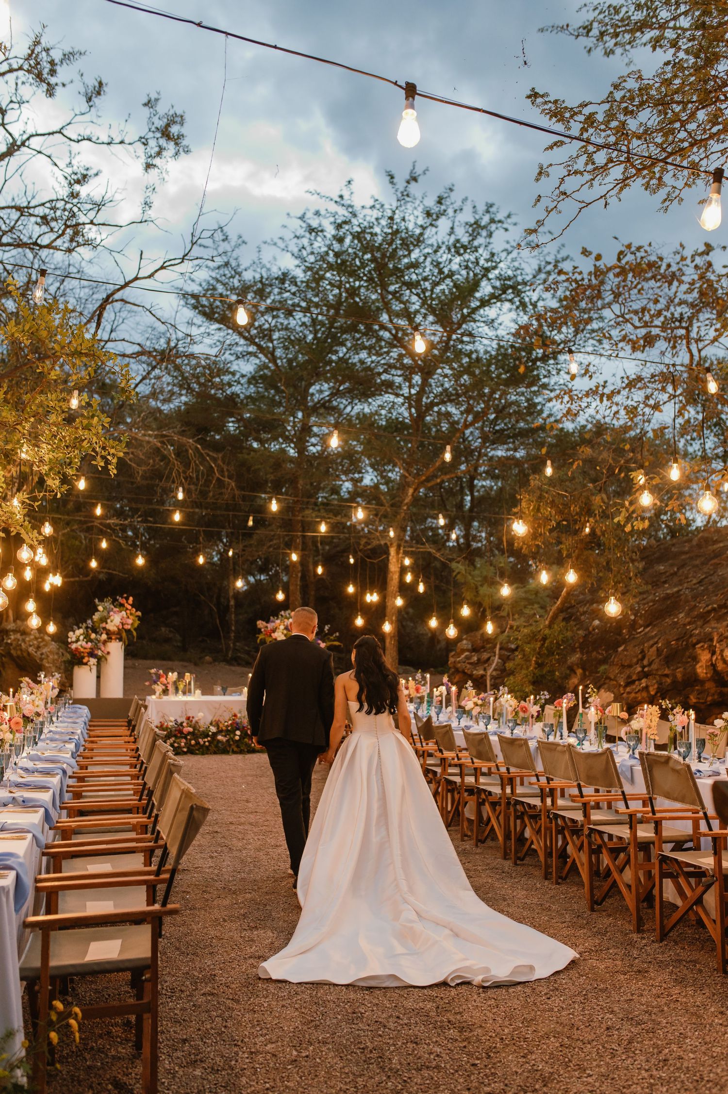 Bride and groom walk hand-in-hand through enchanting outdoor reception venue adorned with Edison bulb lights.