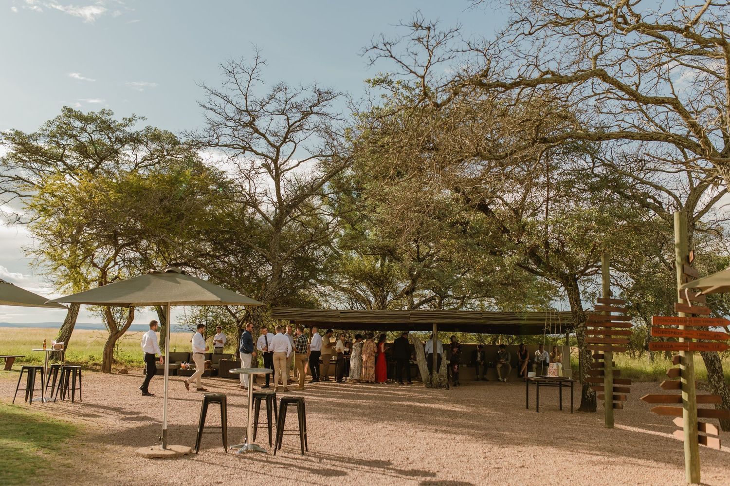 Guests mingling at an outdoor wedding reception under a shaded pavilion surrounded by trees in a natural landscape.