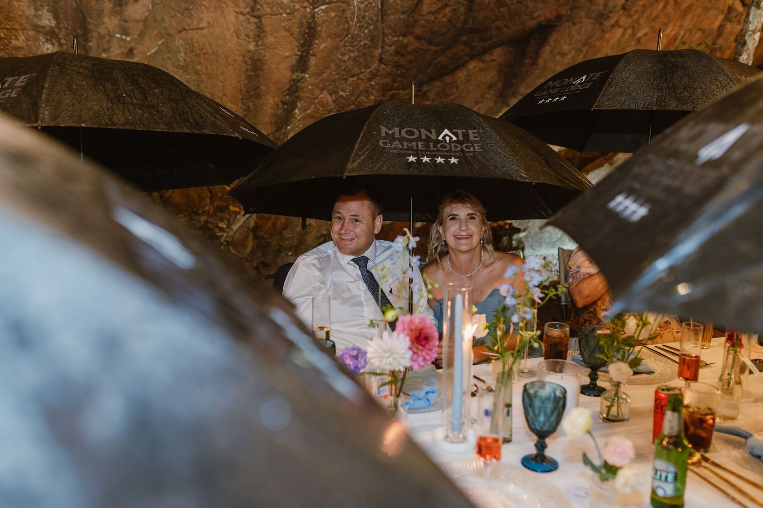 Wedding guests smiling under black Mohale umbrella at elegantly decorated outdoor reception table at night.