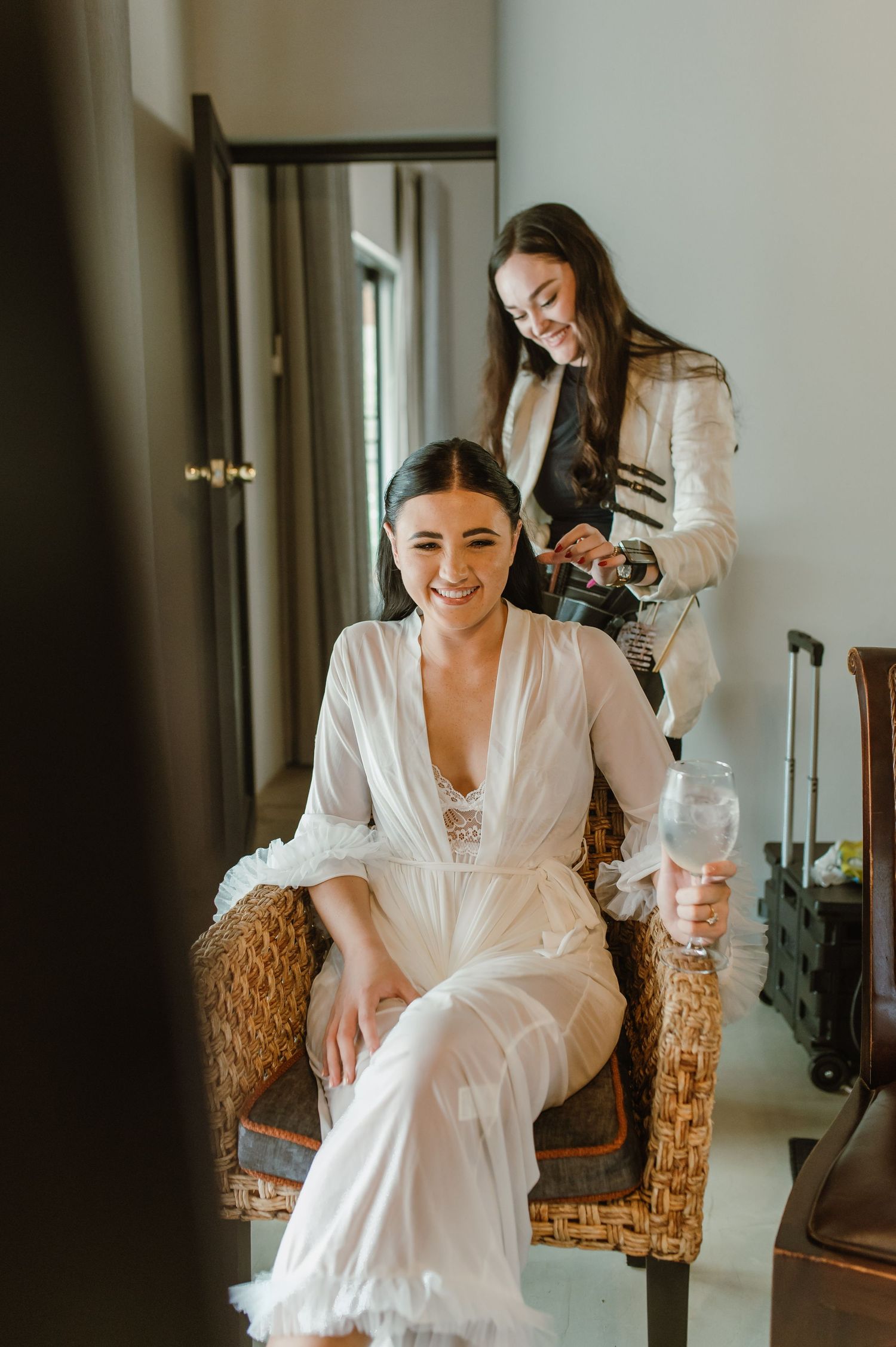 Smiling bride in a white feathered robe sits in a wicker chair while bridesmaid styles her hair.