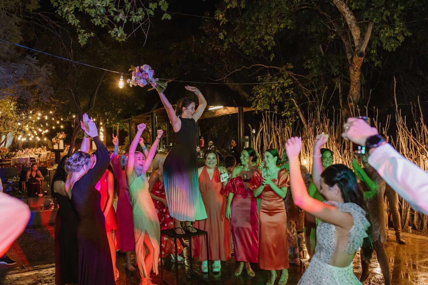 Bride tosses bouquet to excited crowd of guests under string lights at outdoor wedding.