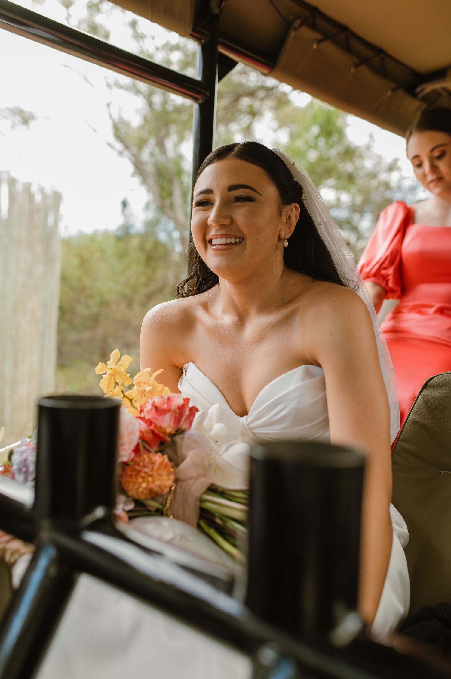 Smiling bride in strapless white gown holding colorful bouquet while riding in a vehicle on her wedding day.