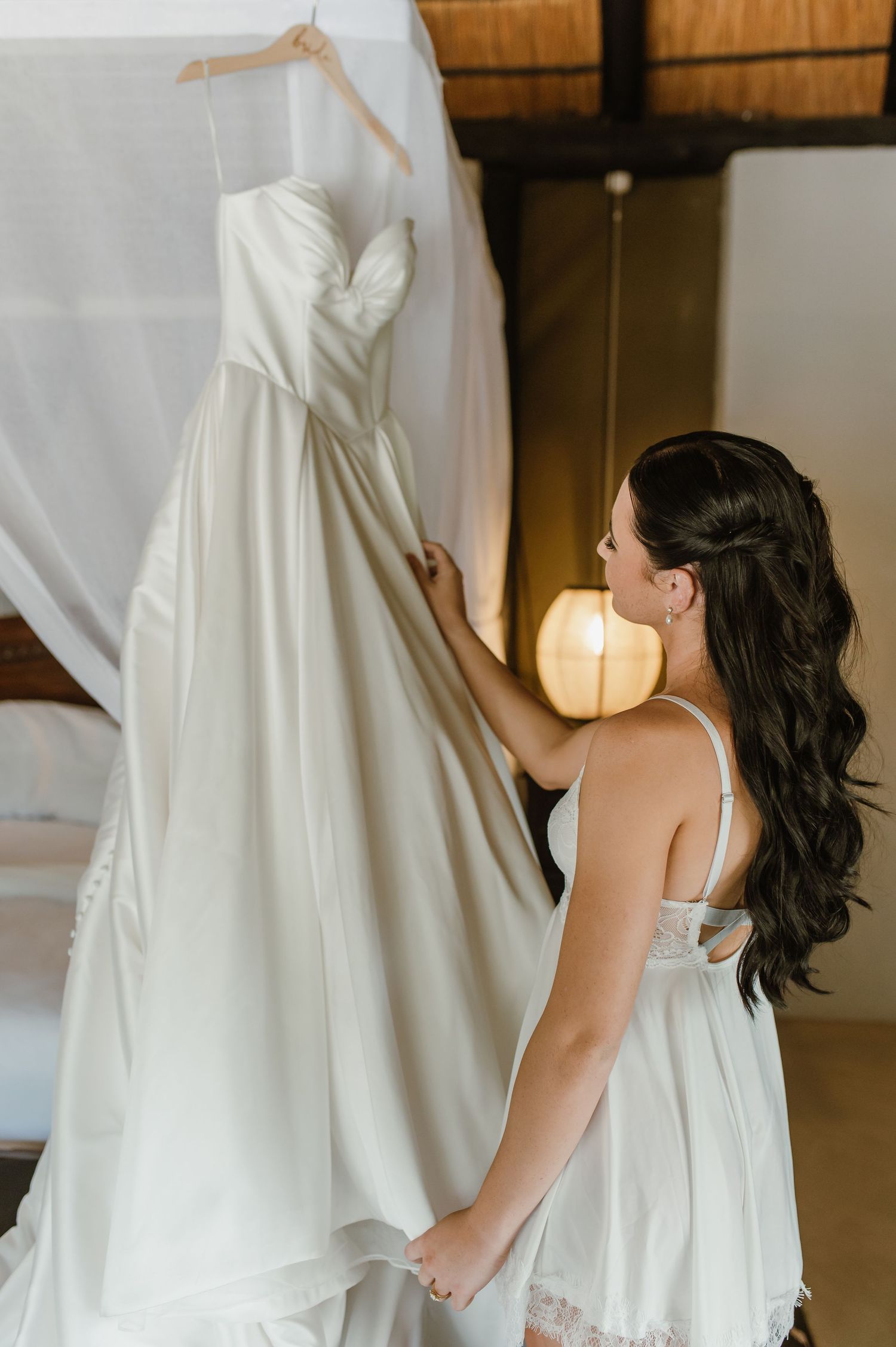 Bride admiring her elegant white wedding gown hanging on a canopy bed frame in a warmly lit room.