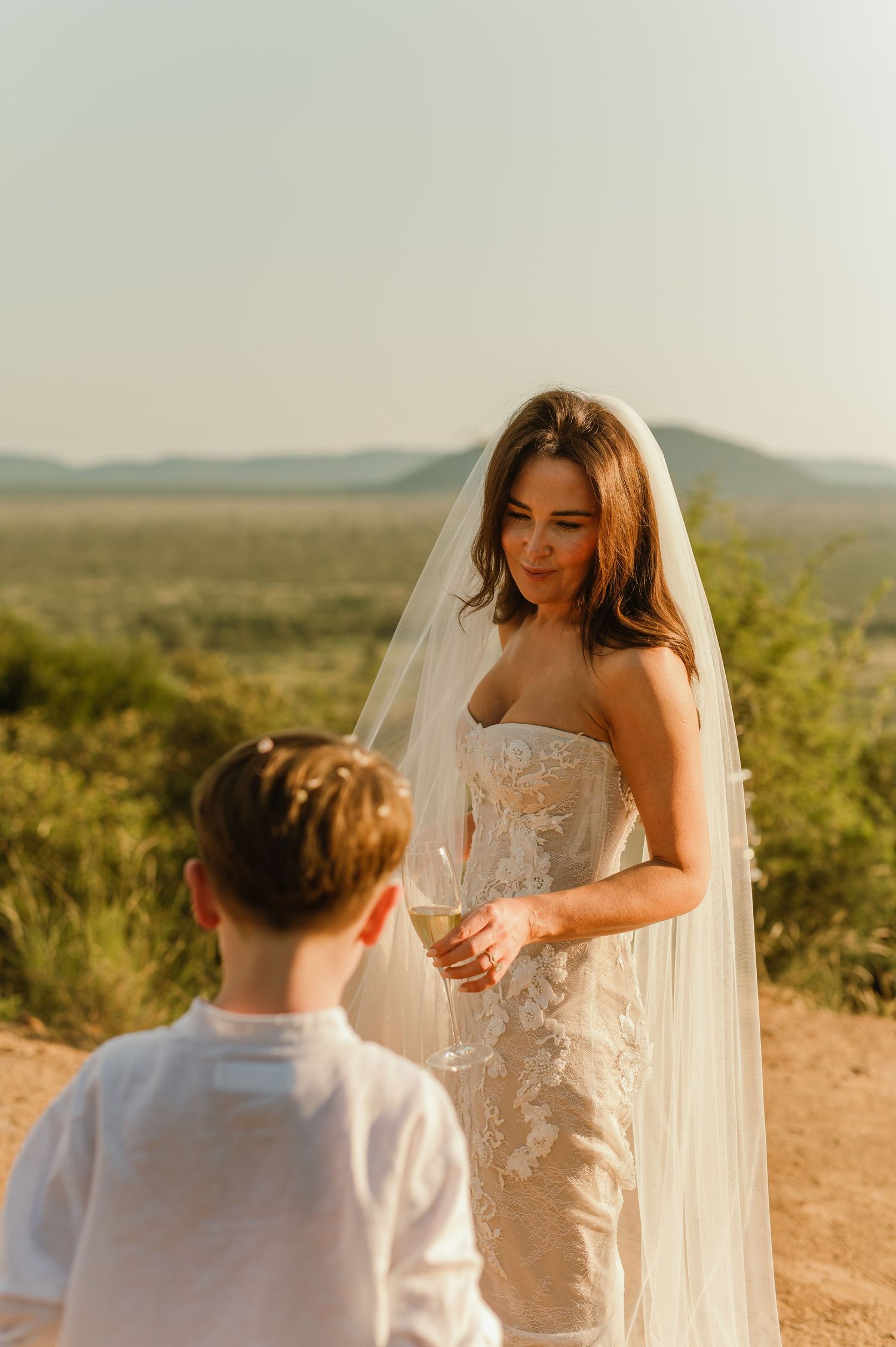 Bride in lace strapless gown and veil shares a moment with young boy at outdoor safari wedding.