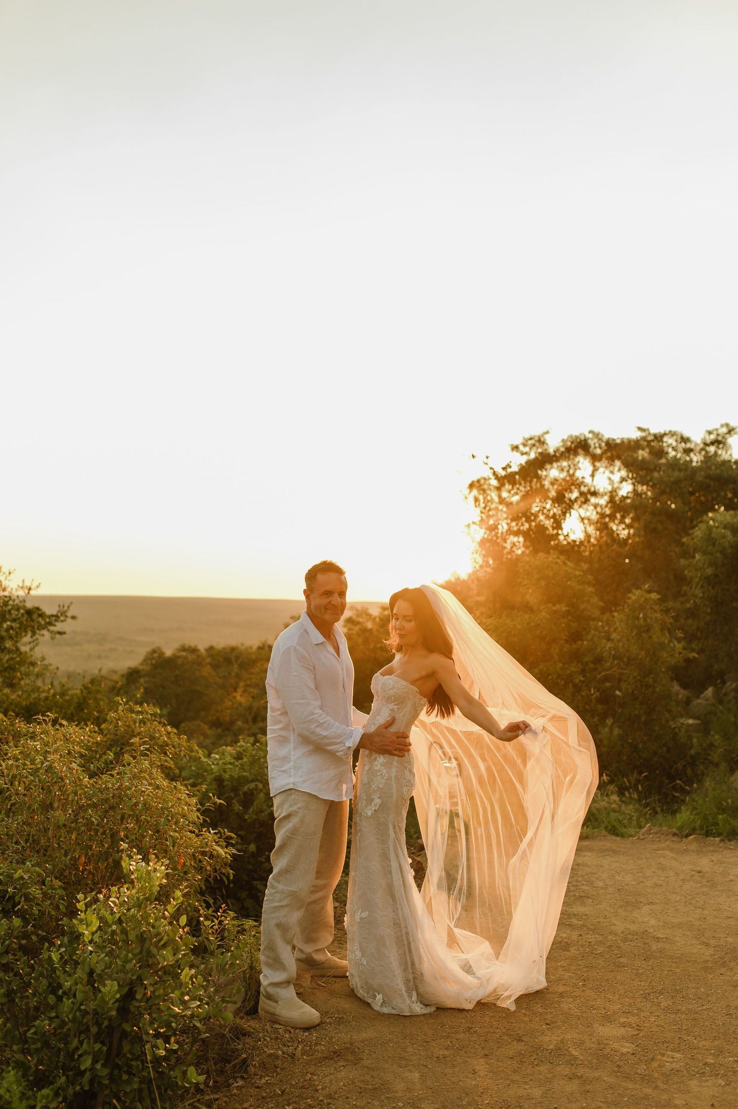 Bride and groom pose at sunset overlook, brides cathedral veil billowing in breeze against golden sky.