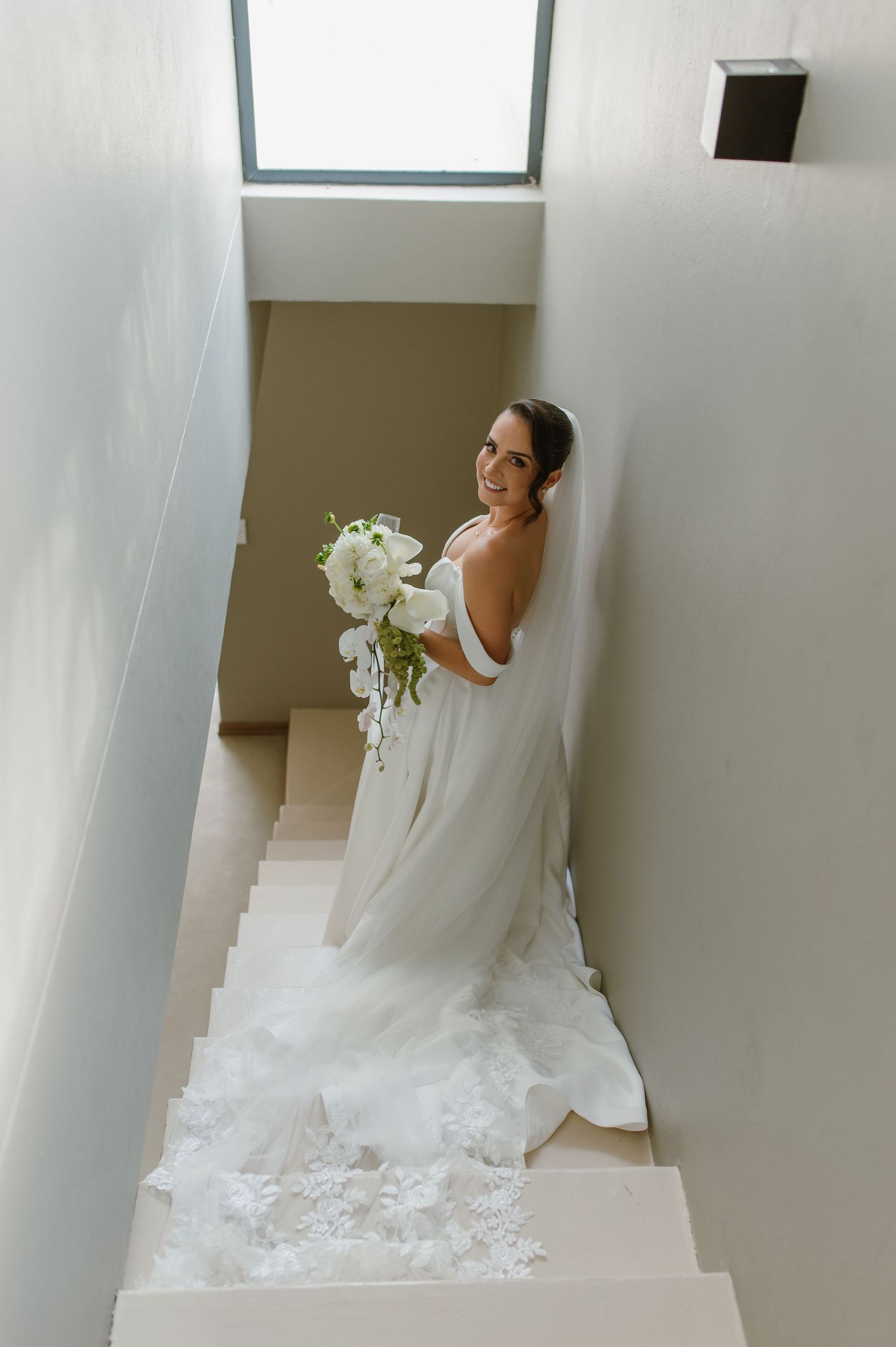 Elegant bride in white gown descending modern white staircase with flowing train.