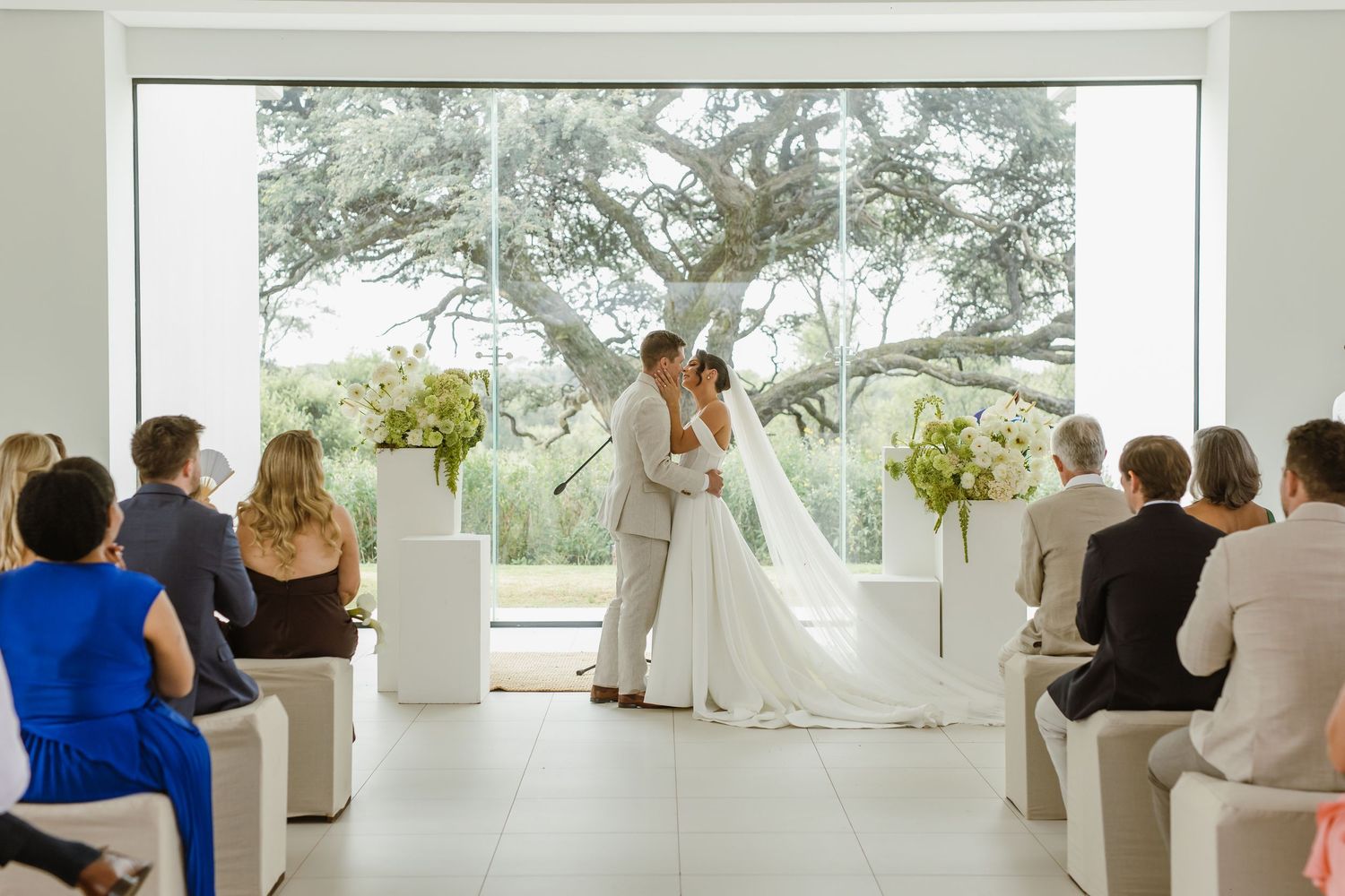 Wedding guests seated in white chairs during an outdoor ceremony with white columns and trees in the background.