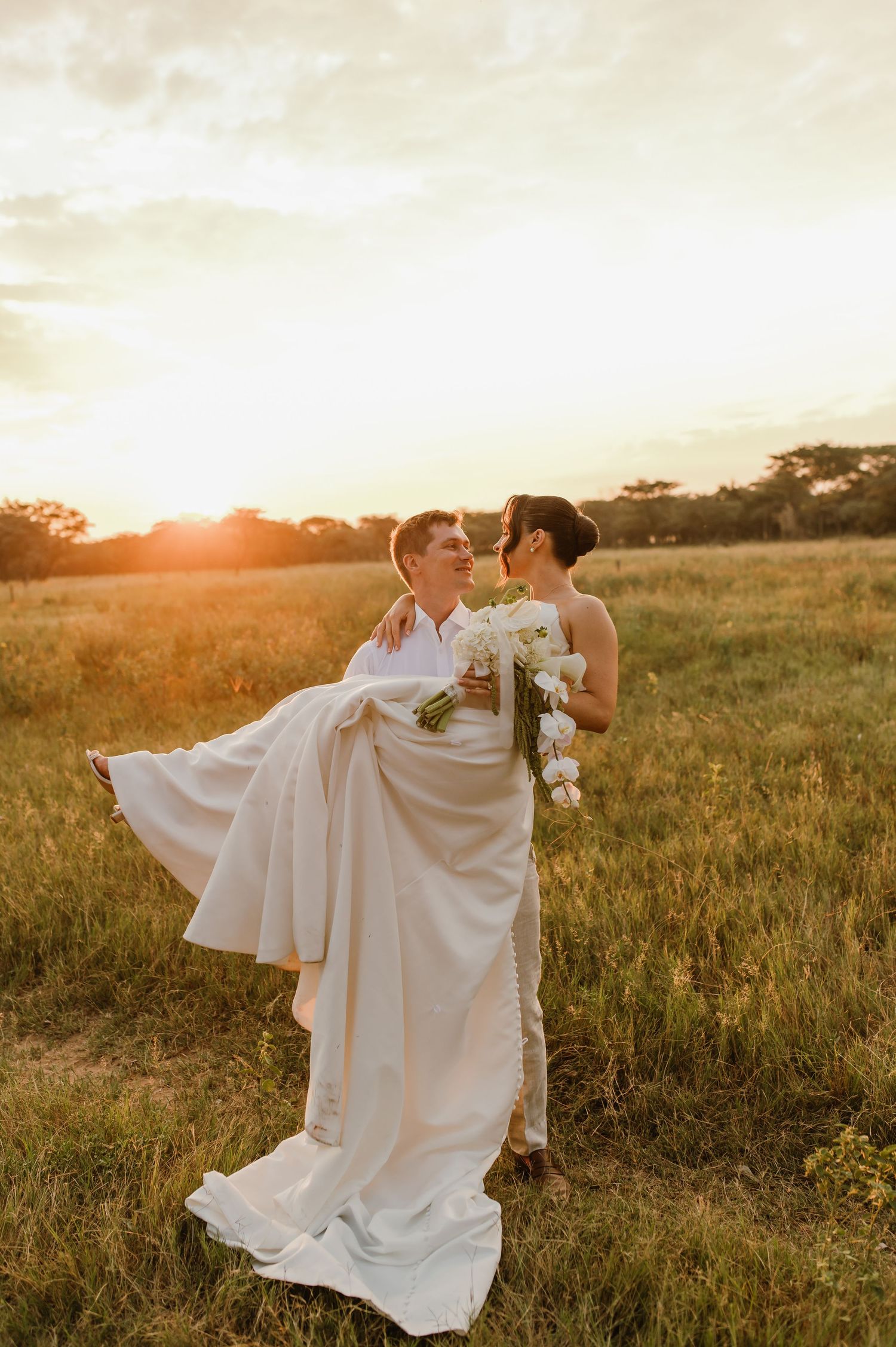 Newlyweds share a romantic moment in a sunlit meadow as the bride's white dress flows in the breeze.
