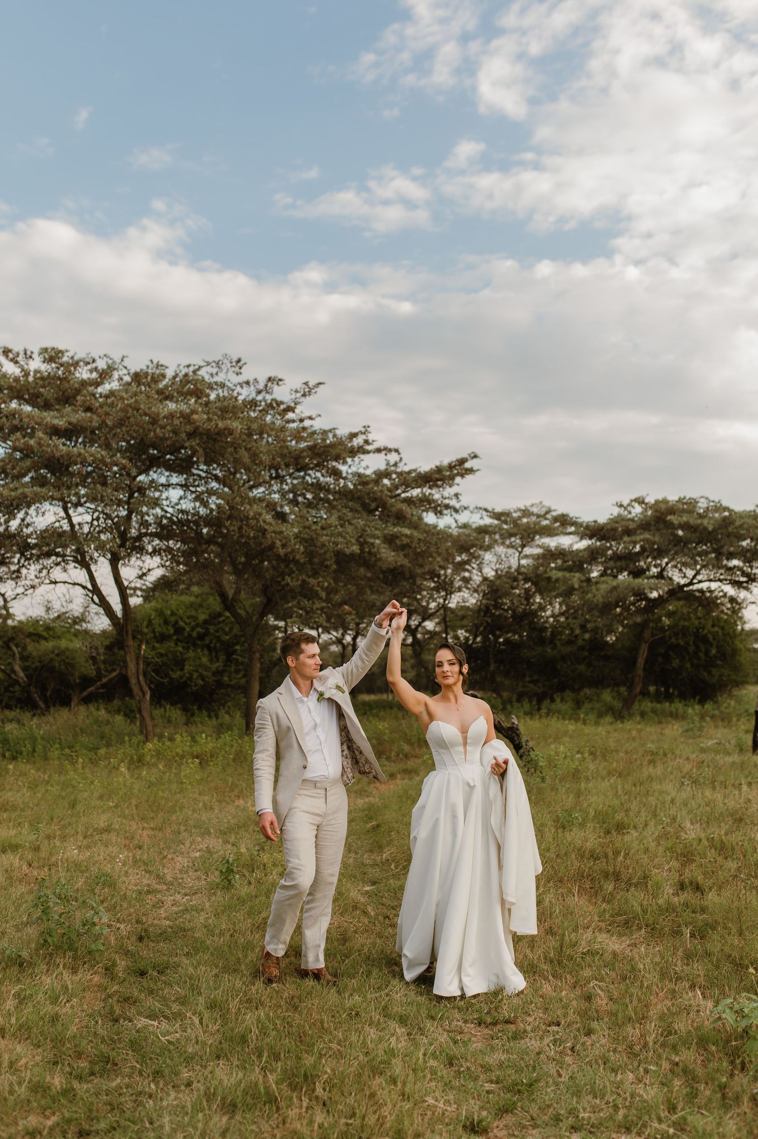 A couple dances together in an open field with acacia trees in the background.