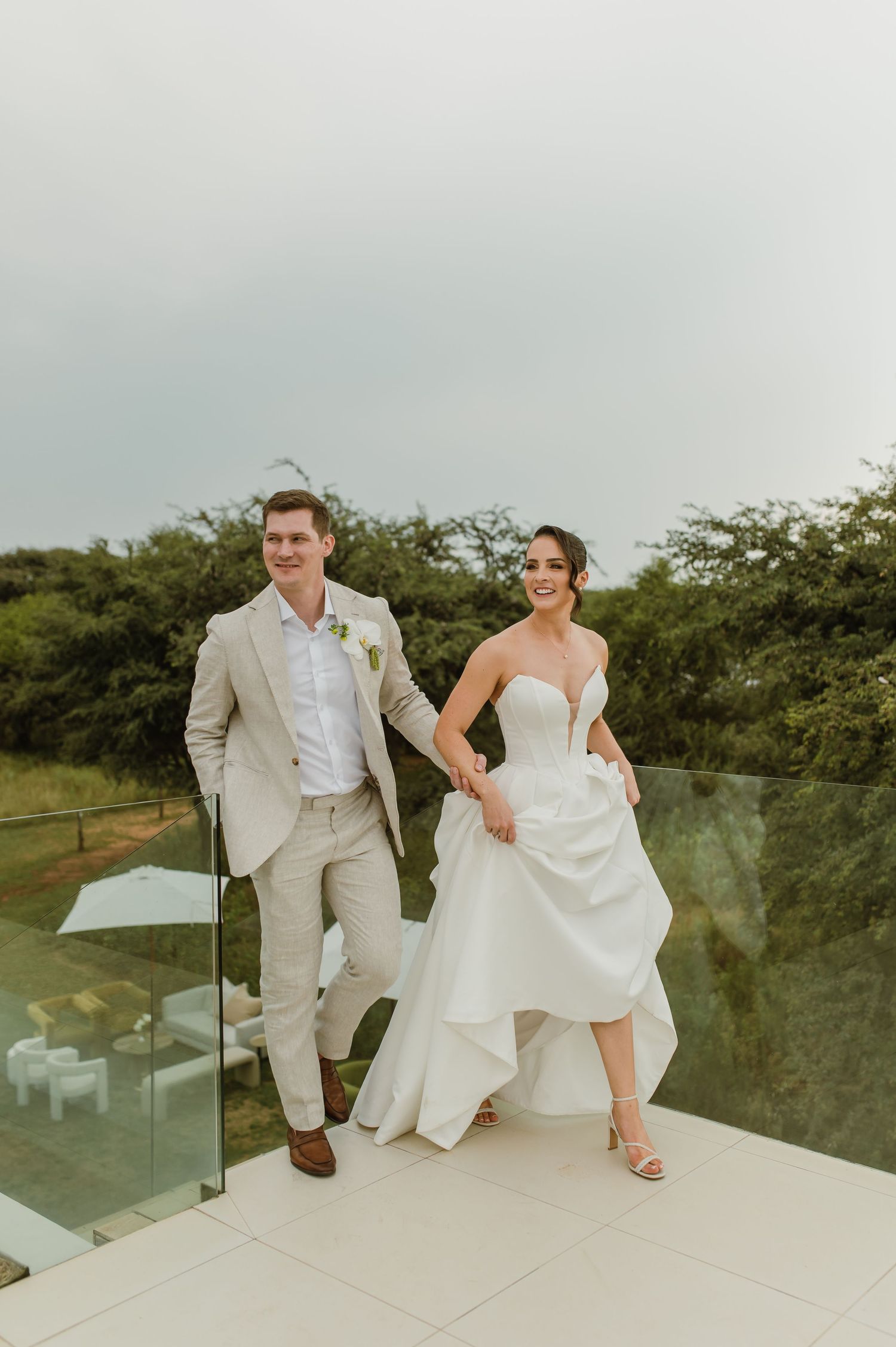 A couple in wedding attire sits casually on a modern glass-railed balcony overlooking green scenery.