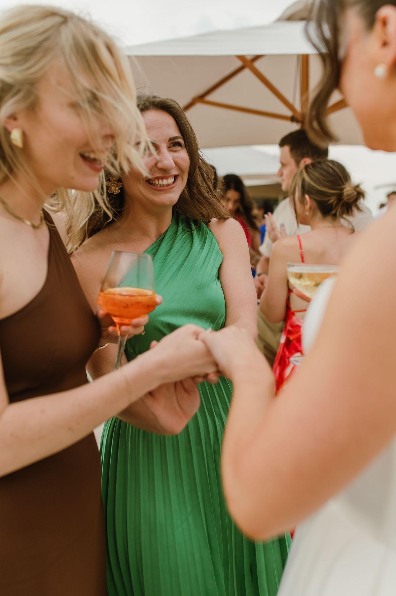 Friends in colorful dresses laugh and celebrate at an outdoor party under white umbrellas.