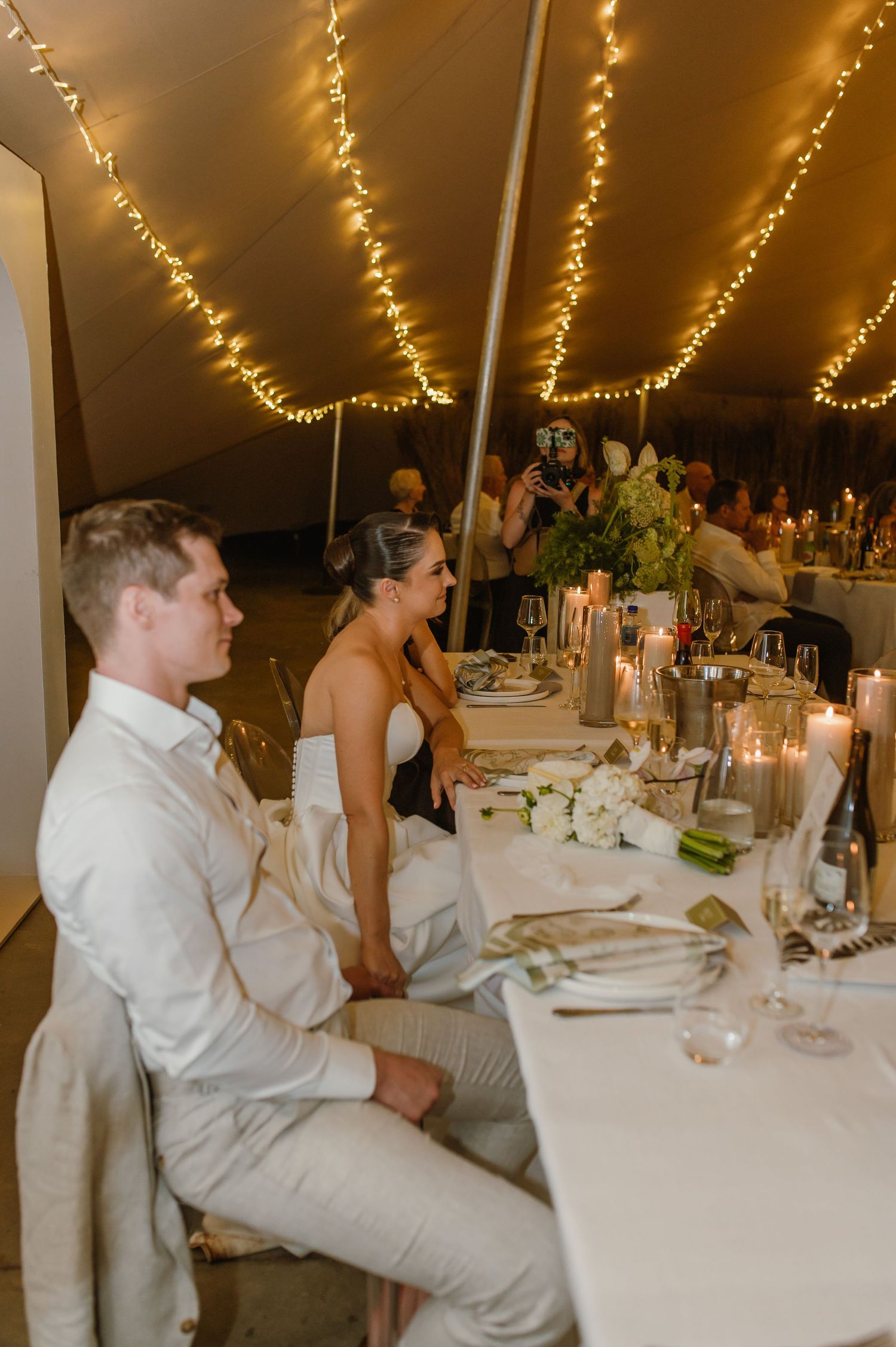 Wedding guests seated at elegant tables under twinkling lights in a reception tent.