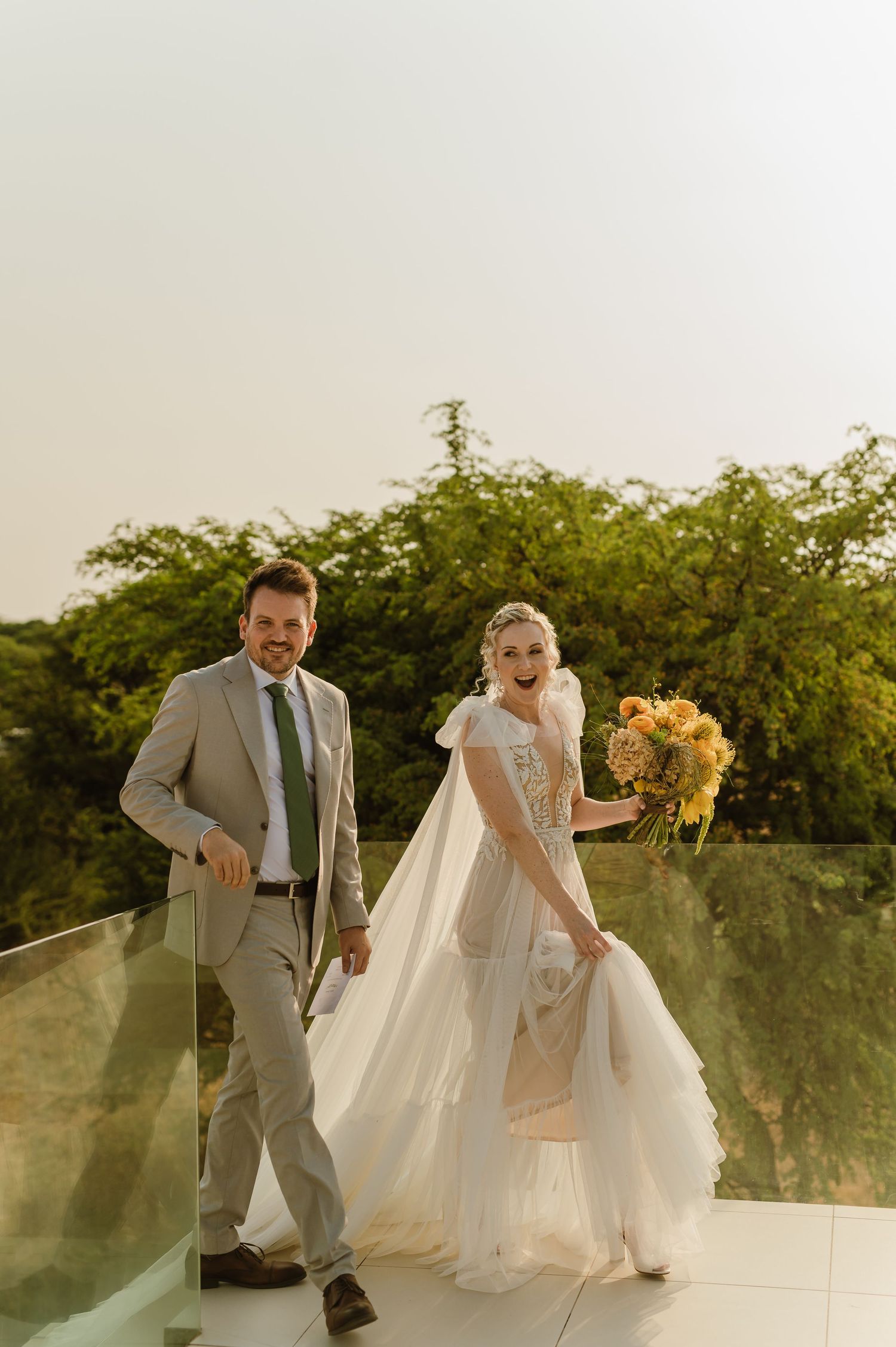 Couple poses on a terrace with yellow bouquet during golden hour wedding photography.
