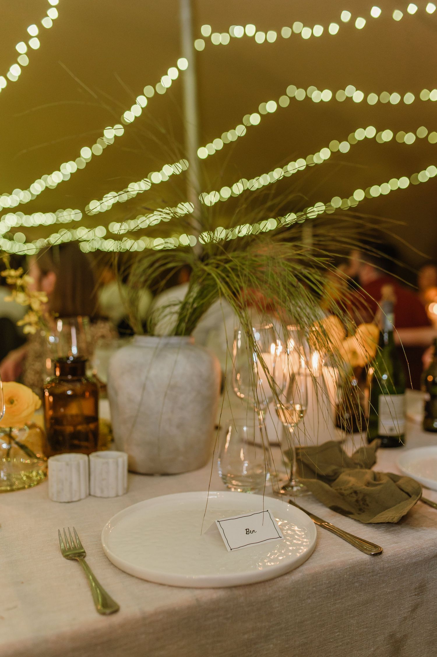 Minimalist table settings with glassware and floral accents under string light canopy.