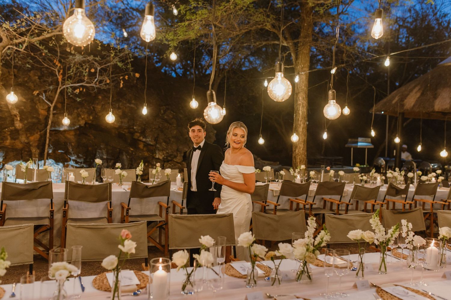 Long banquet tables set for an outdoor wedding reception with hanging Edison bulbs creating ambient lighting.