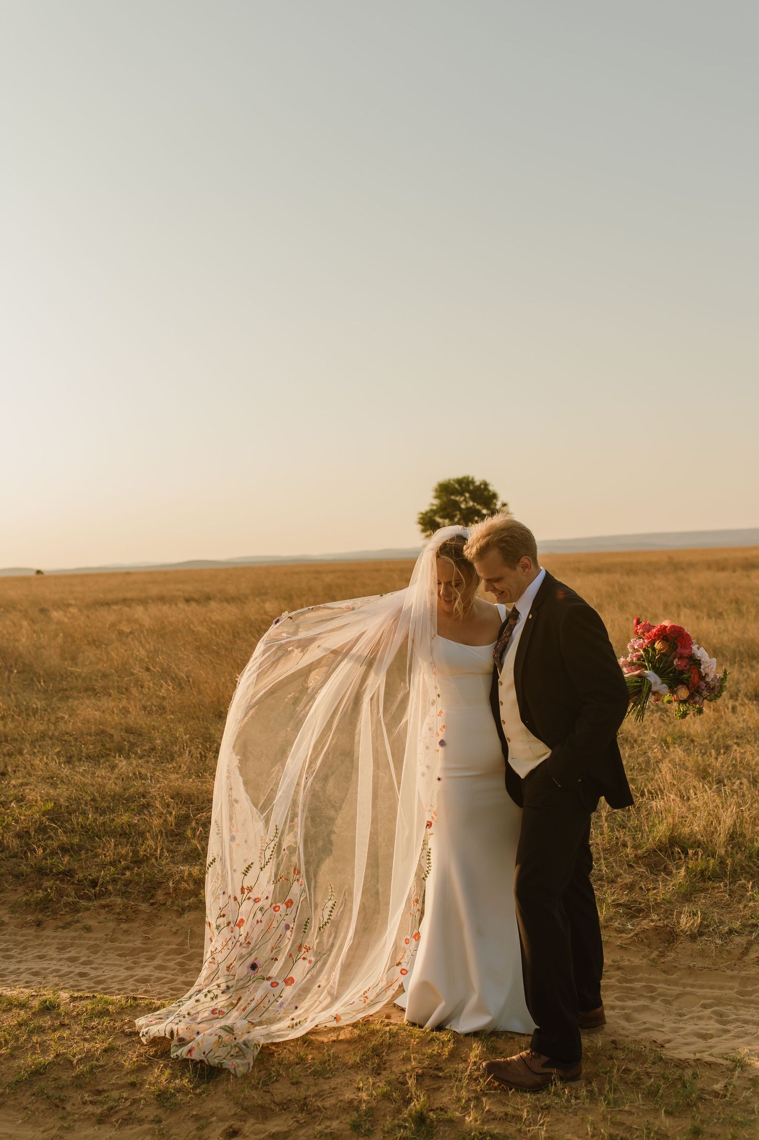 A couple embraces during their sunset wedding photos as a long veil flows in the breeze.