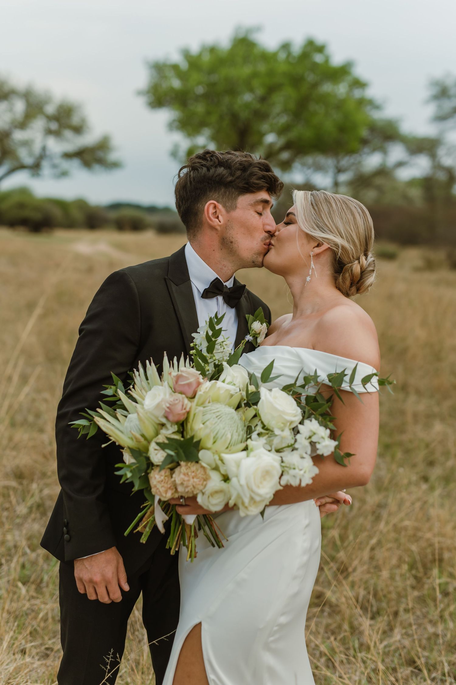 Romantic wedding couple kisses while holding elegant white bouquet in natural grassland setting.