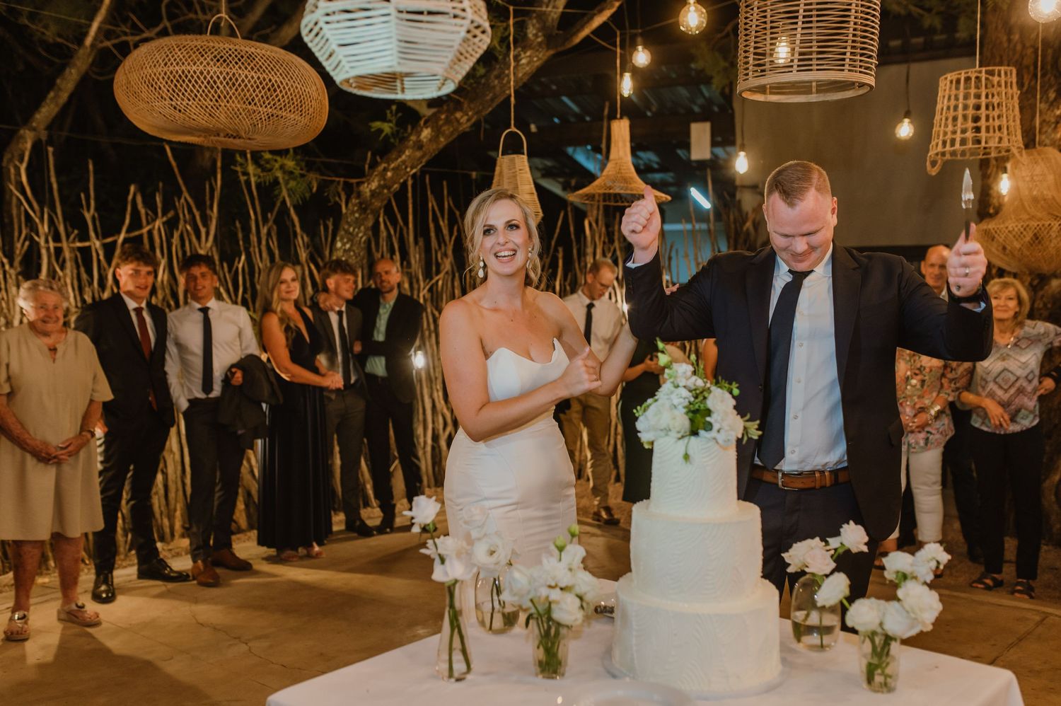 Wedding celebration with guests gathered around a white tiered cake under ambient string lights.