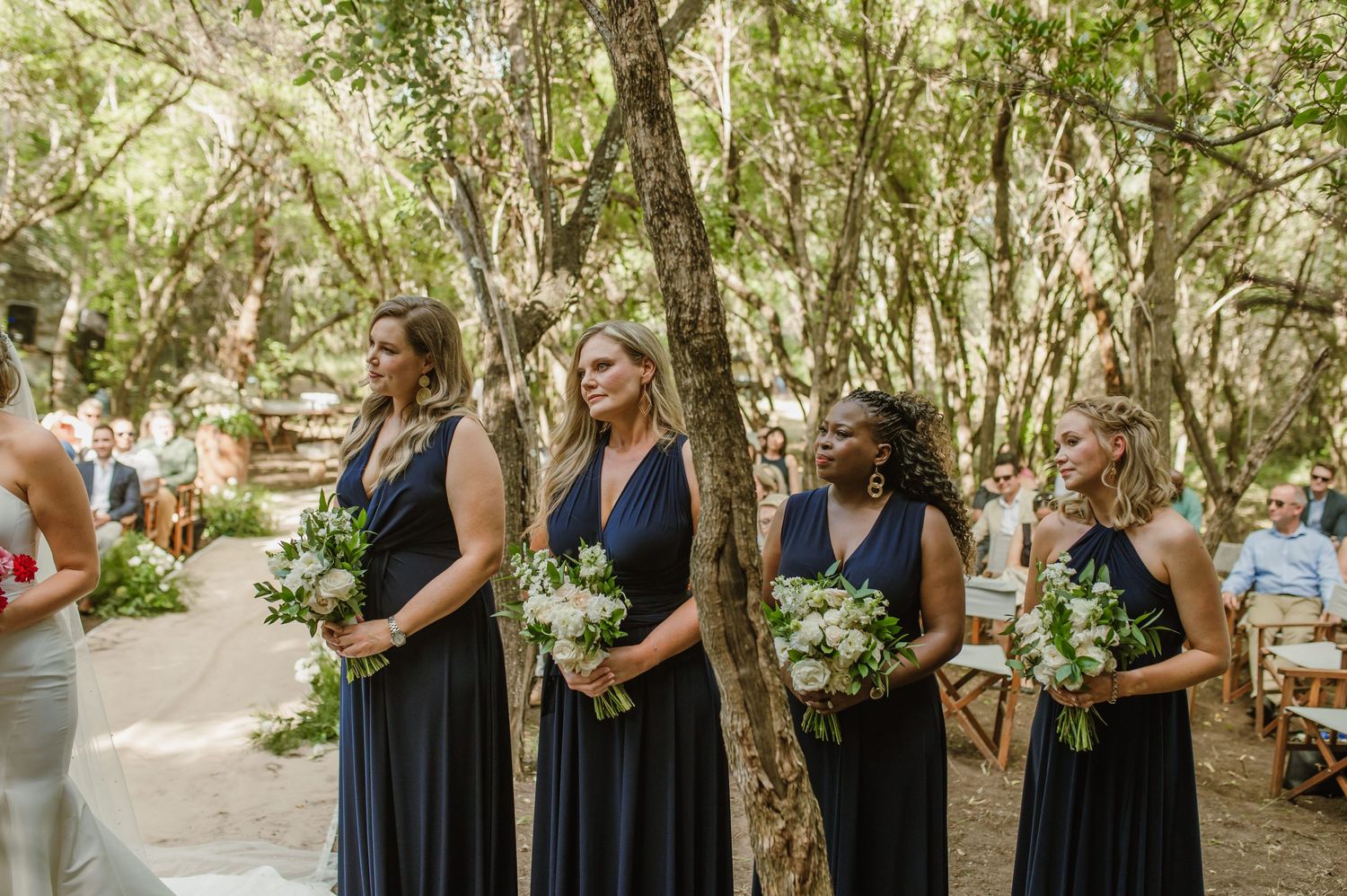 Bridesmaids in navy dresses stand in line during outdoor forest wedding ceremony.
