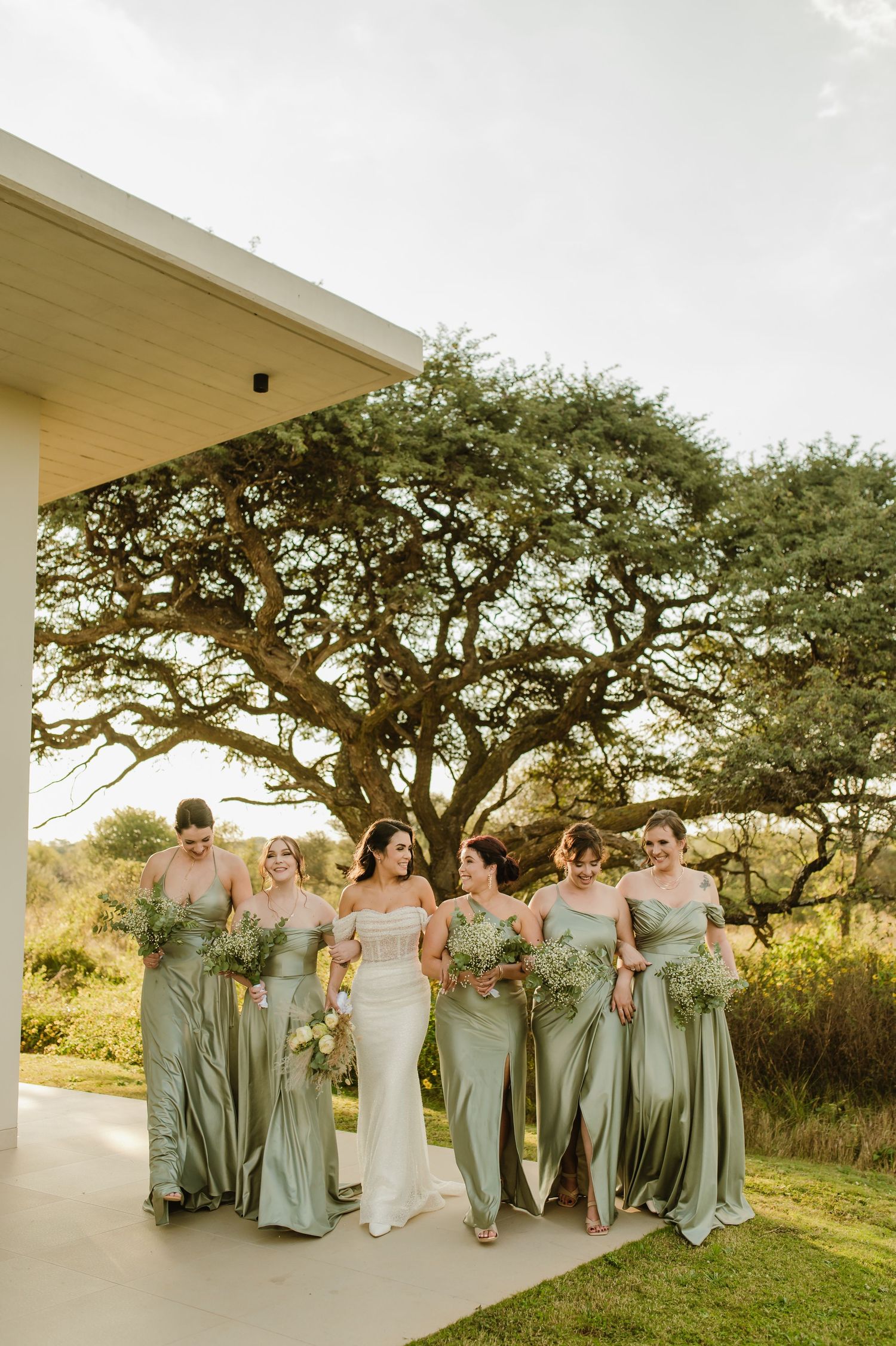 Wedding party in sage green dresses posed together on a covered porch with trees in background.