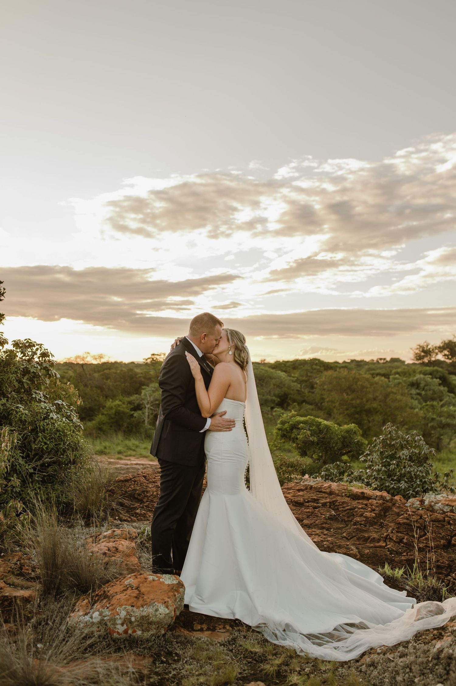 Romantic sunset portrait of newlyweds embracing on rocky hilltop with flowing white dress.