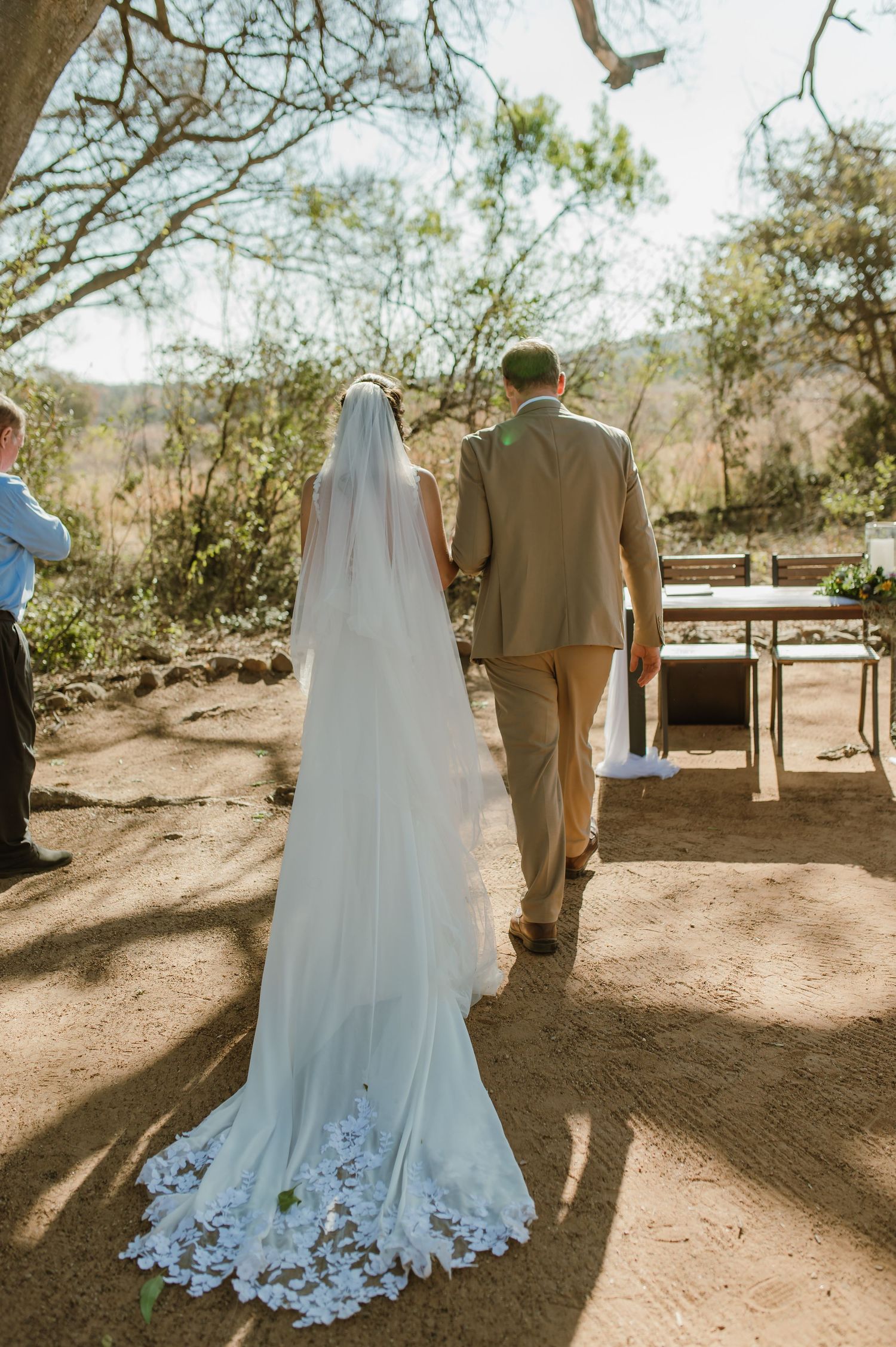 Bride in flowing white dress and groom walk together on desert path under bare trees.