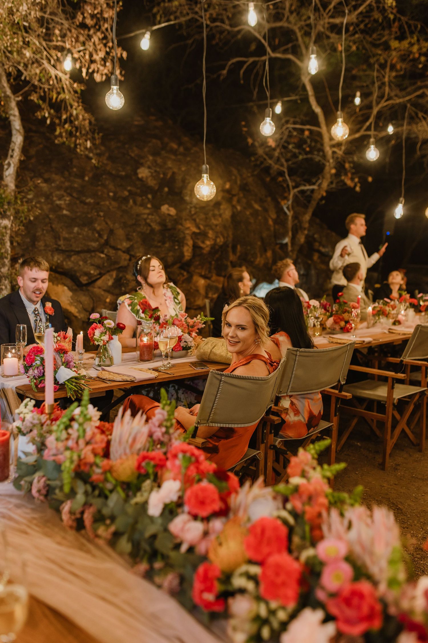 Long wooden tables decorated with pink and red floral arrangements under string lights at outdoor reception.