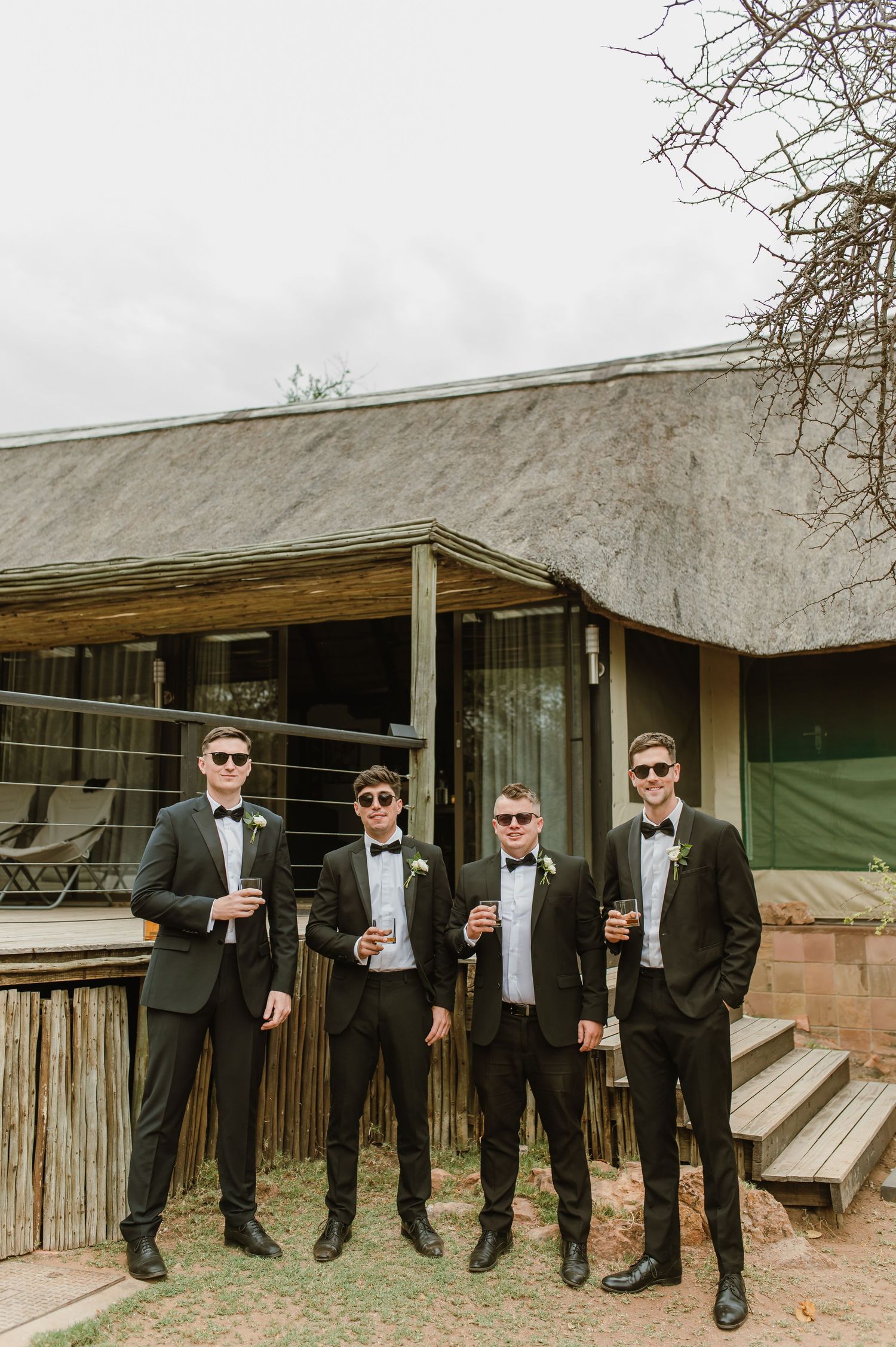 Groomsmen lined up against rustic wooden fence wearing matching black tuxedos and sunglasses.