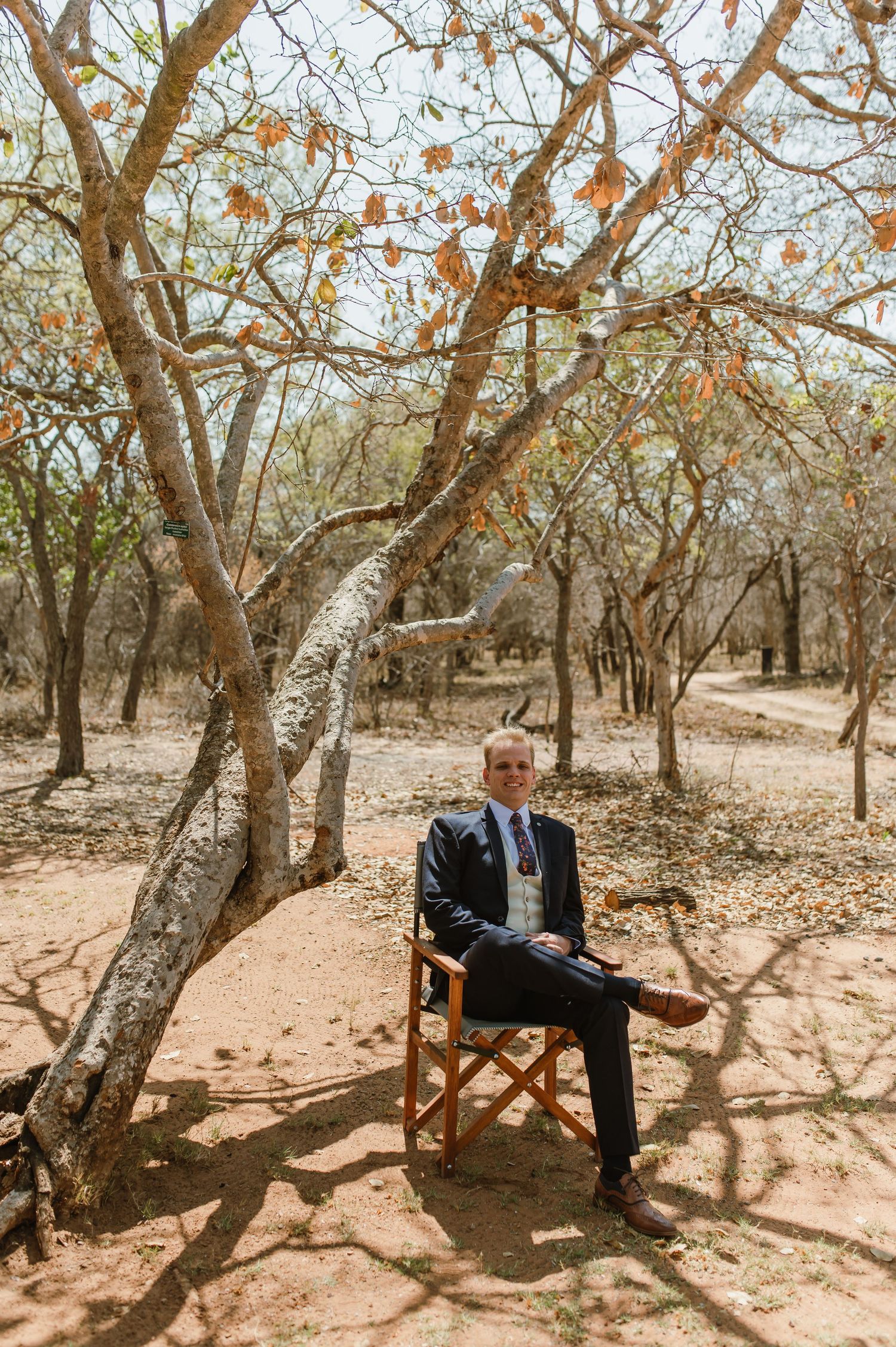 A suited figure sits on a wooden chair beneath autumn trees in a rustic outdoor setting.