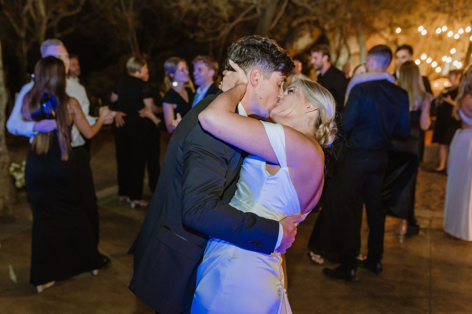 Couples slow dancing at wedding reception under romantic outdoor lighting.