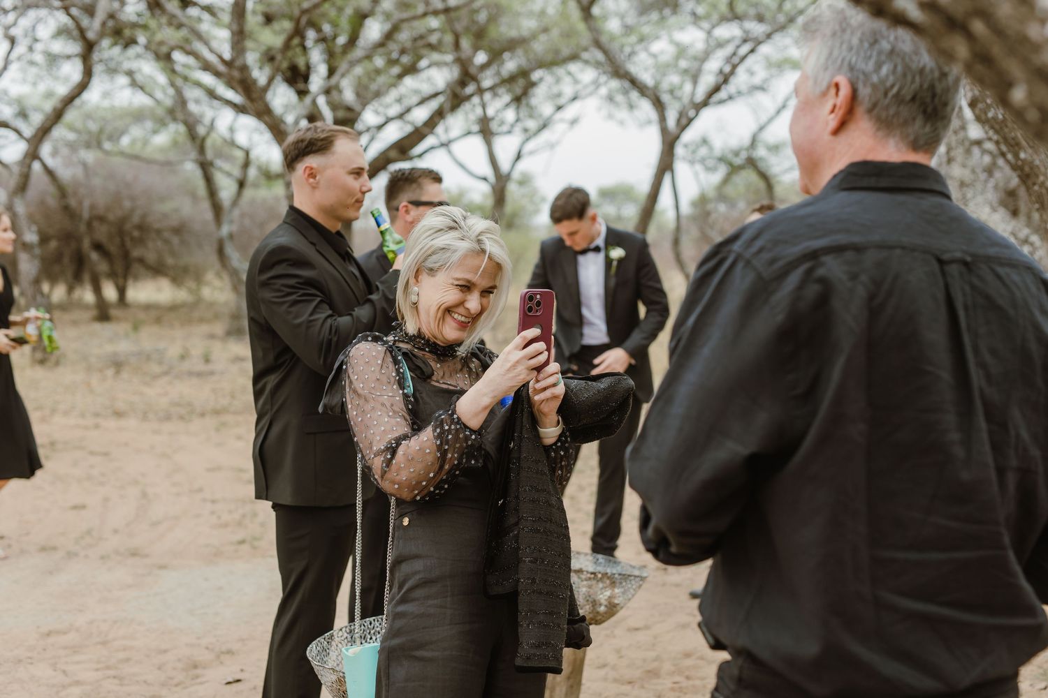 Wedding guests in black attire gather outdoors under trees in a rustic setting.