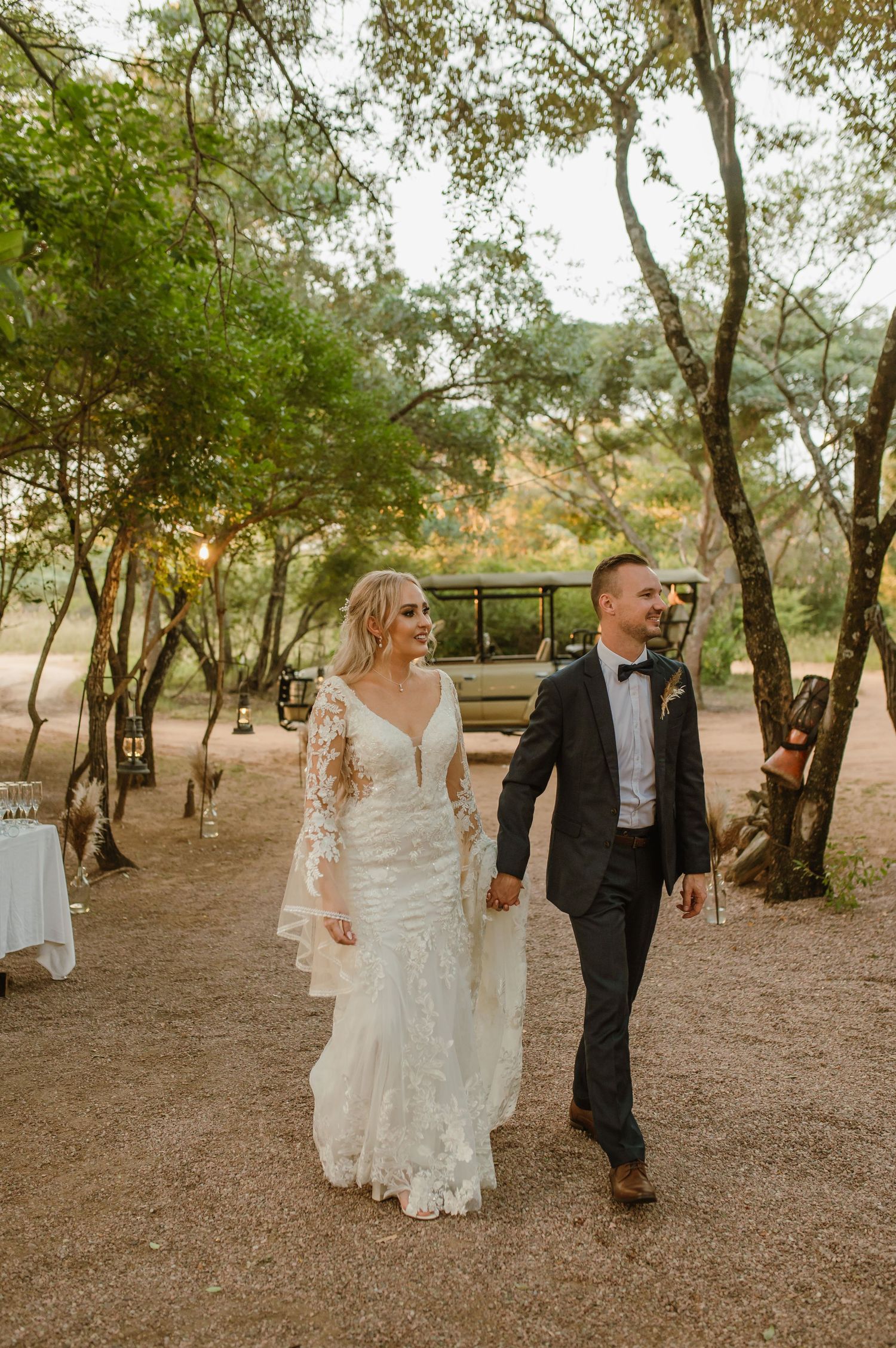 A couple walks hand in hand through a wooded path in elegant wedding attire.