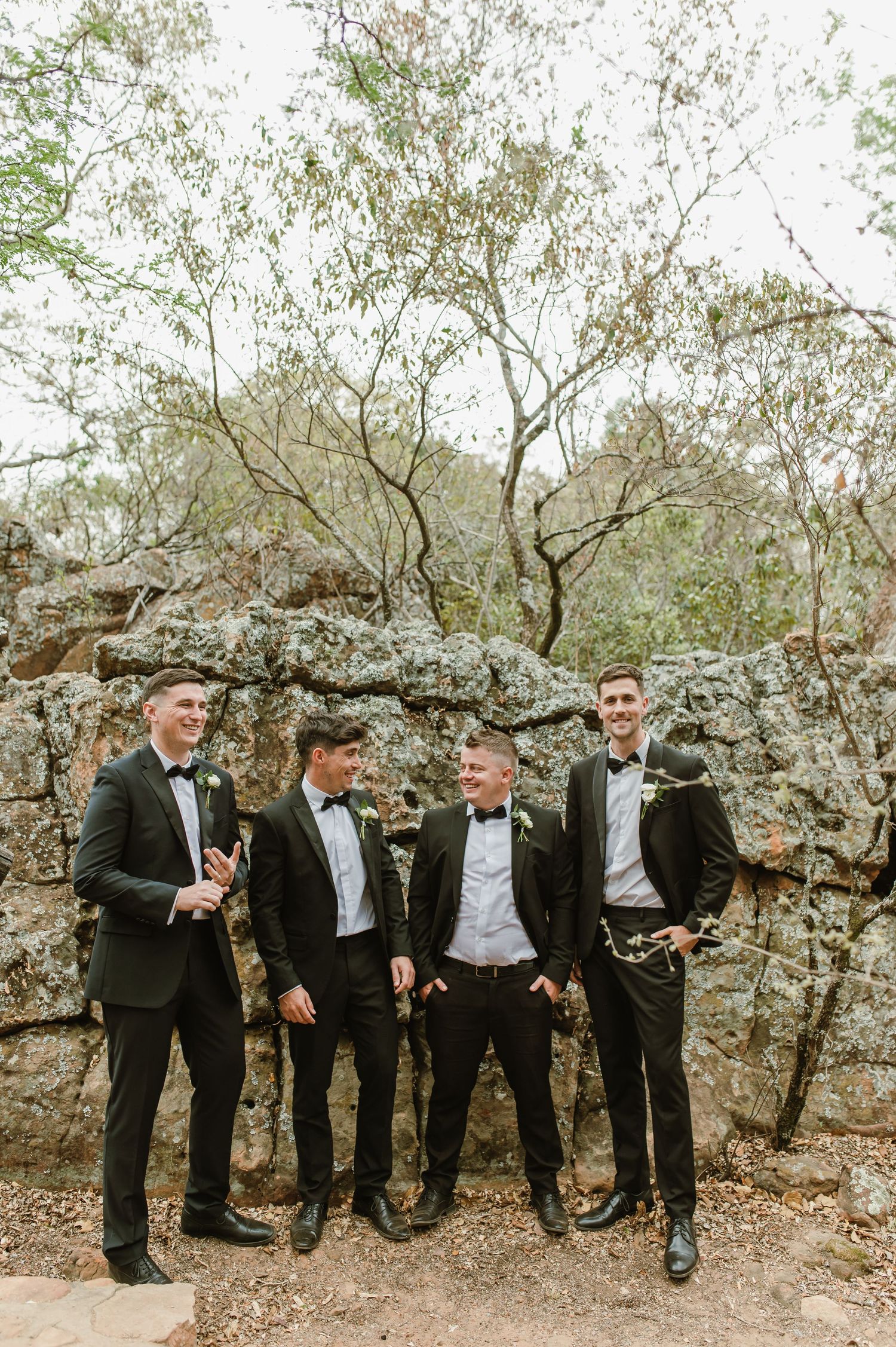 Group of groomsmen in black tuxedos laugh and interact against a rocky natural backdrop.