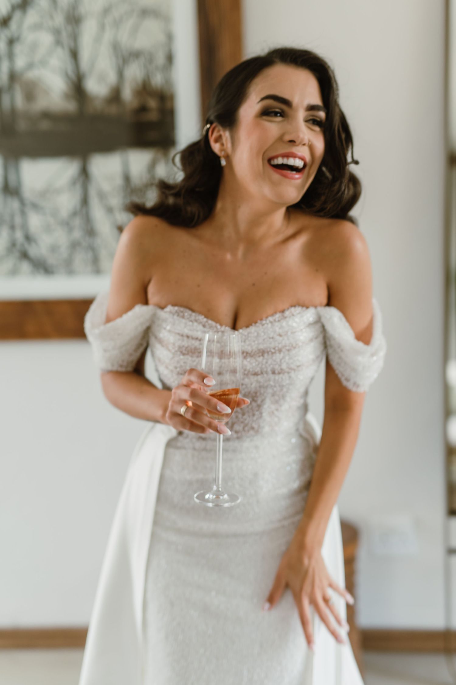 Bride in a detailed white off-shoulder gown laughs while holding a champagne glass.