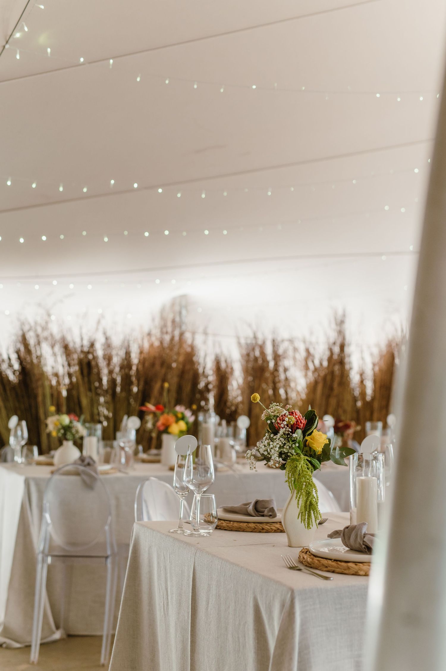 Elegant wedding reception table setup with white linens, string lights, and floral centerpieces in a rustic venue.