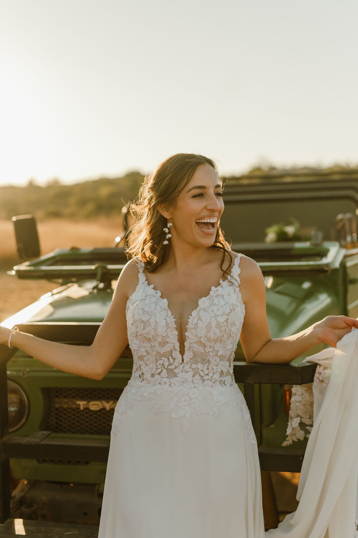 A person in a white lace wedding dress laughs joyfully next to a black vehicle at sunset.