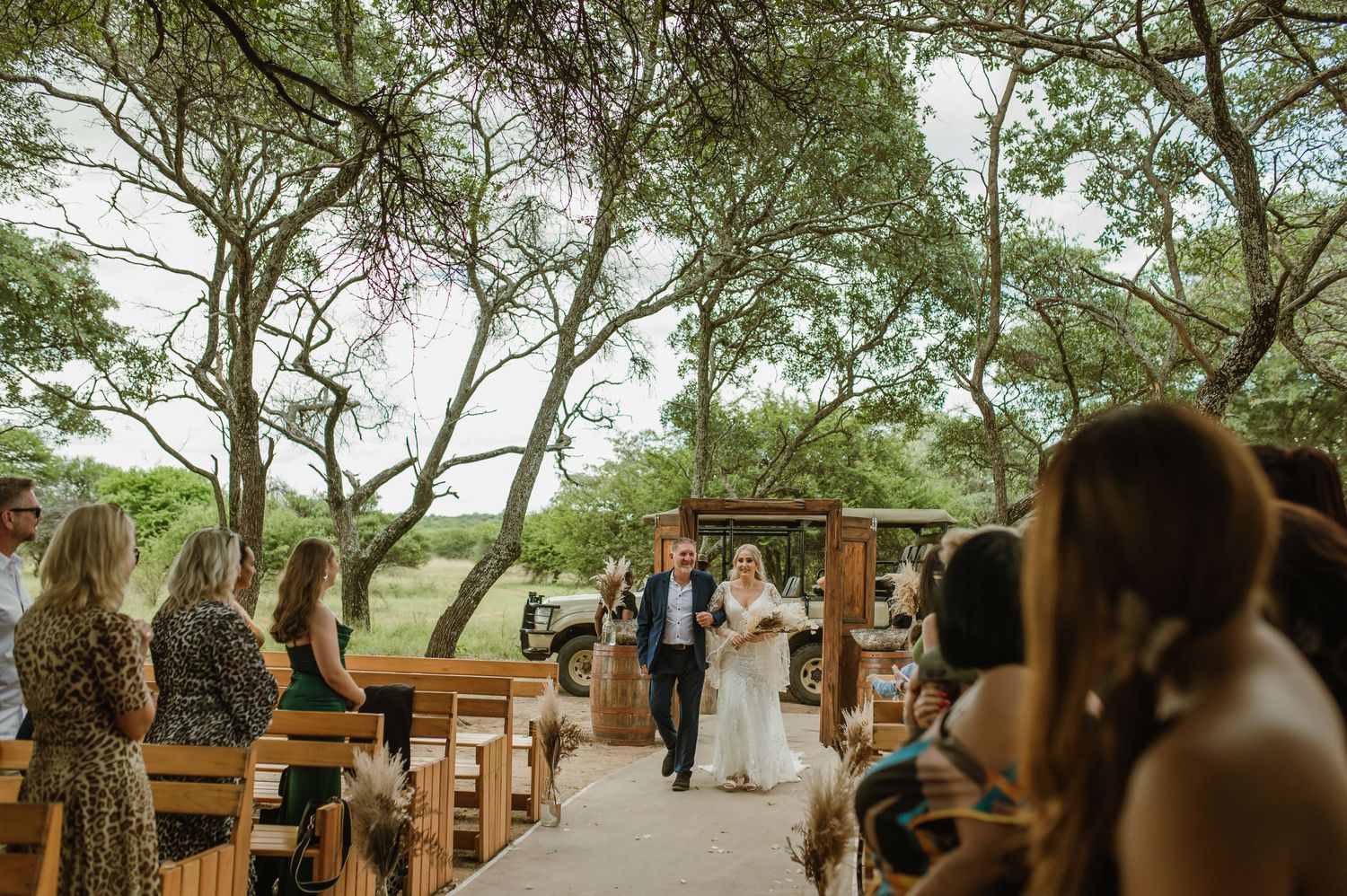 Wooden benches arranged in rows beneath shady trees for an outdoor wedding ceremony.
